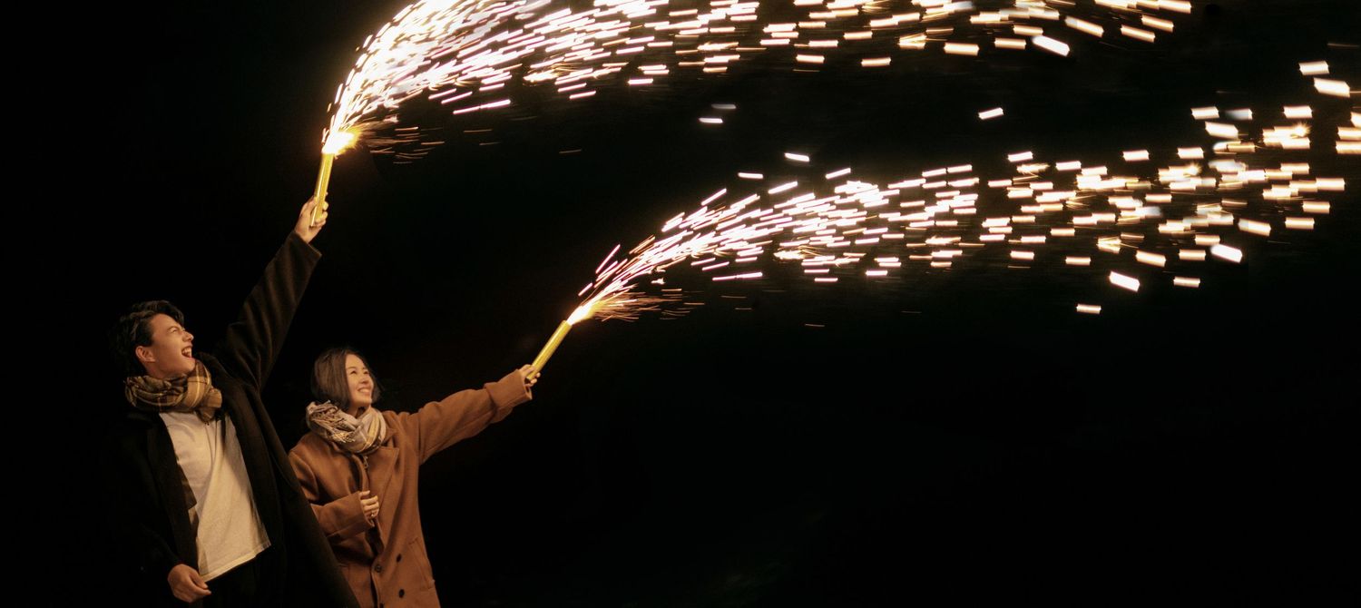 Two people holding sparkling fireworks against a dark night sky creating streaks of light.