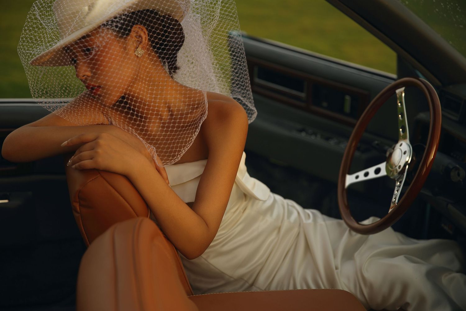 Bride in white dress and veiled hat resting inside a vintage convertible car interior.