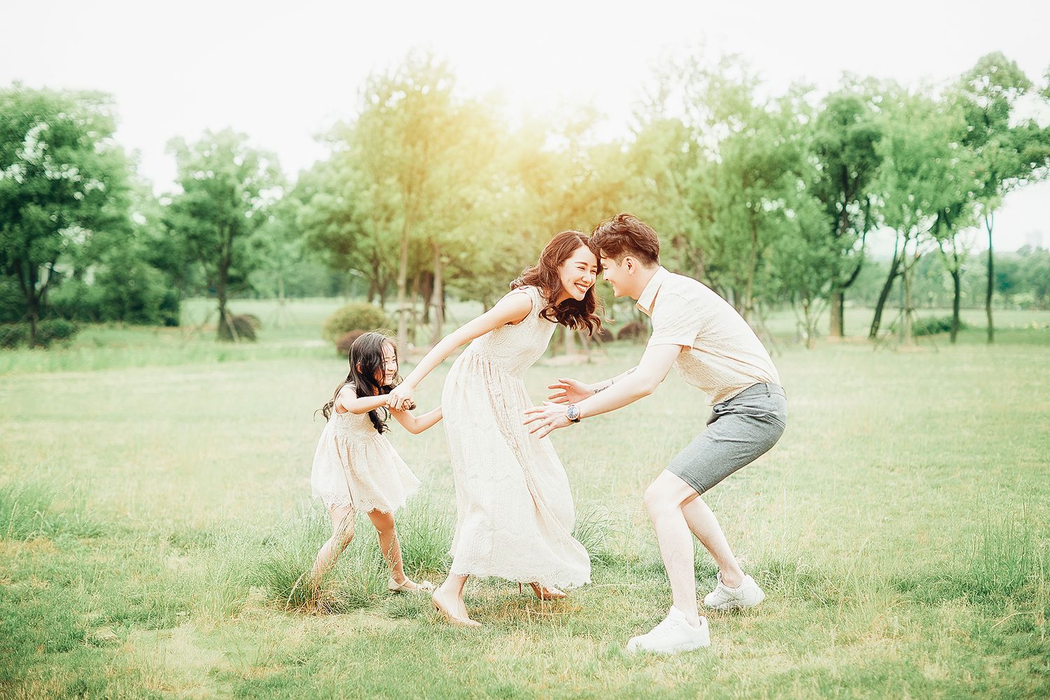 A joyful Asian family plays together in a sunlit park, with parents and daughter dancing on grass.
