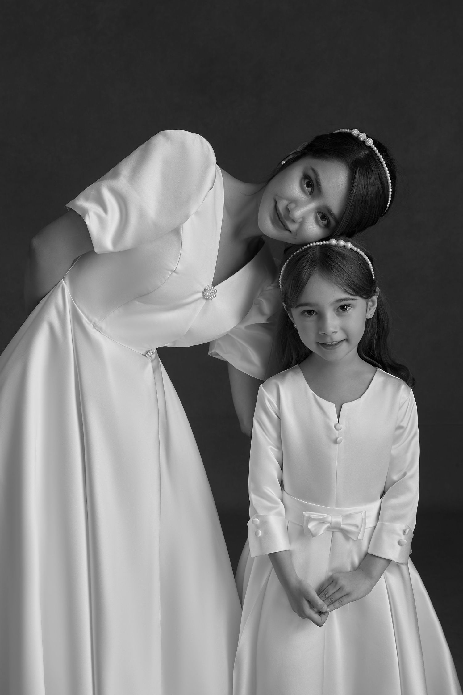 Black and white photo of a bride in white gown bending down next to a young girl in a white dress with bow.