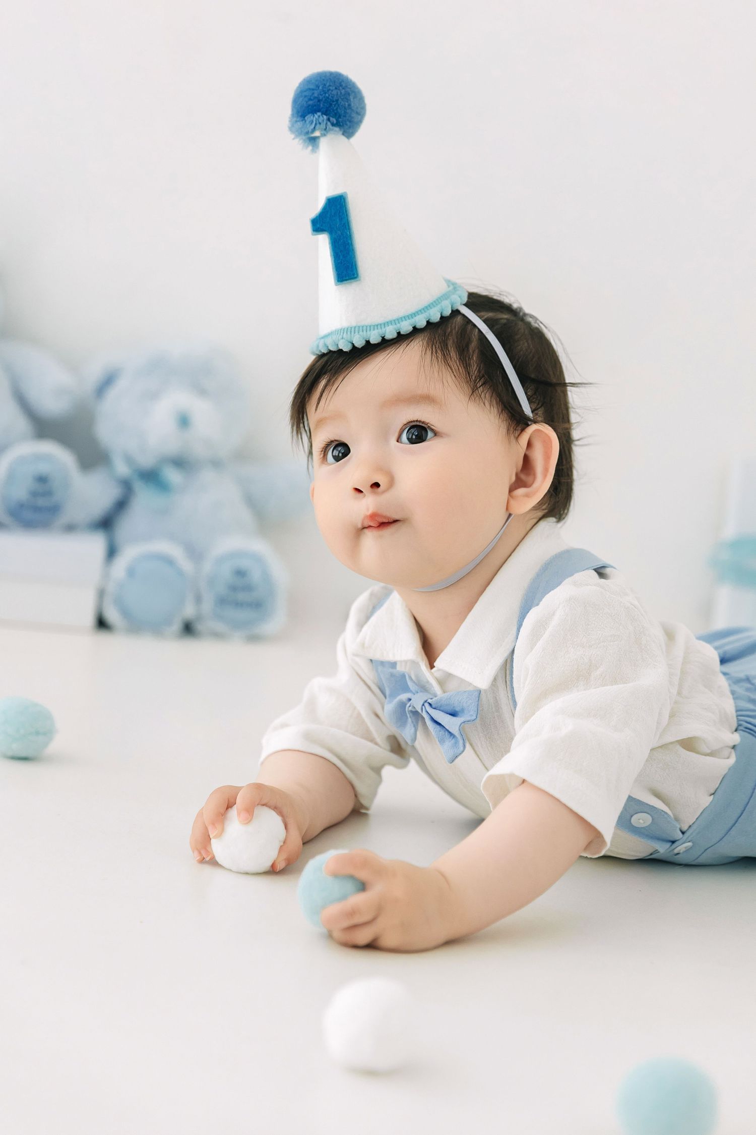 A baby celebrates their first birthday wearing a blue party hat and bow tie outfit, surrounded by blue toys and balls.