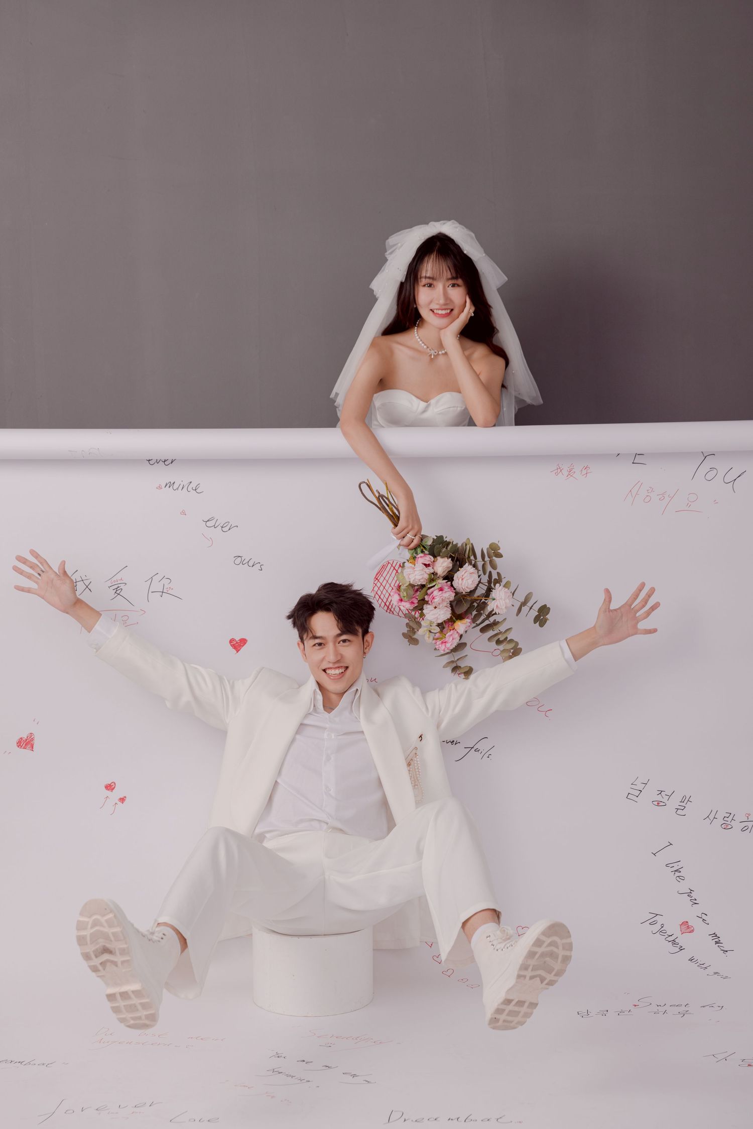 A playful wedding photo with the groom seated arms wide open and the bride peeking over a white backdrop.