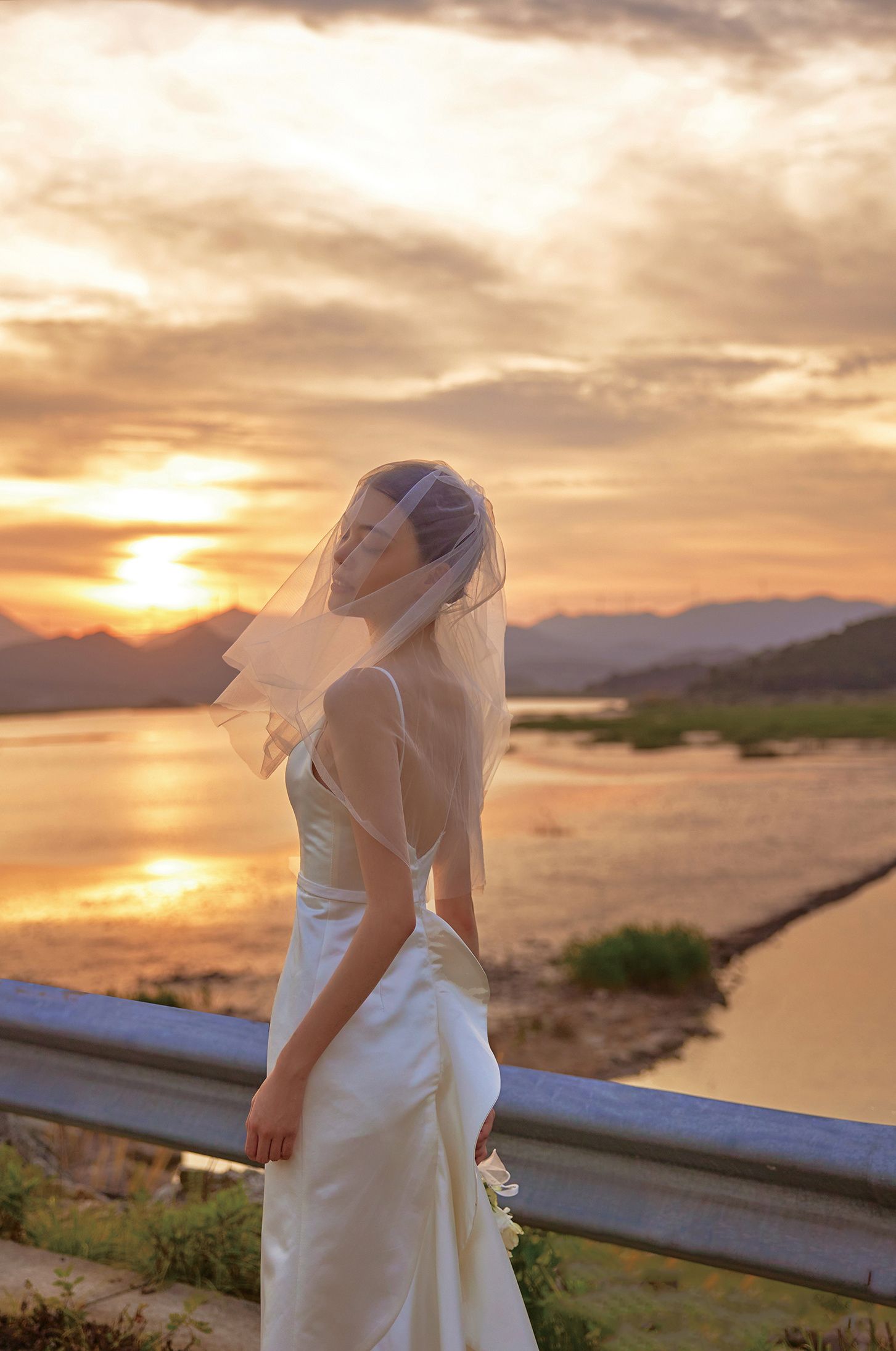 Bride in white gown and veil poses at sunset overlooking a serene lake.