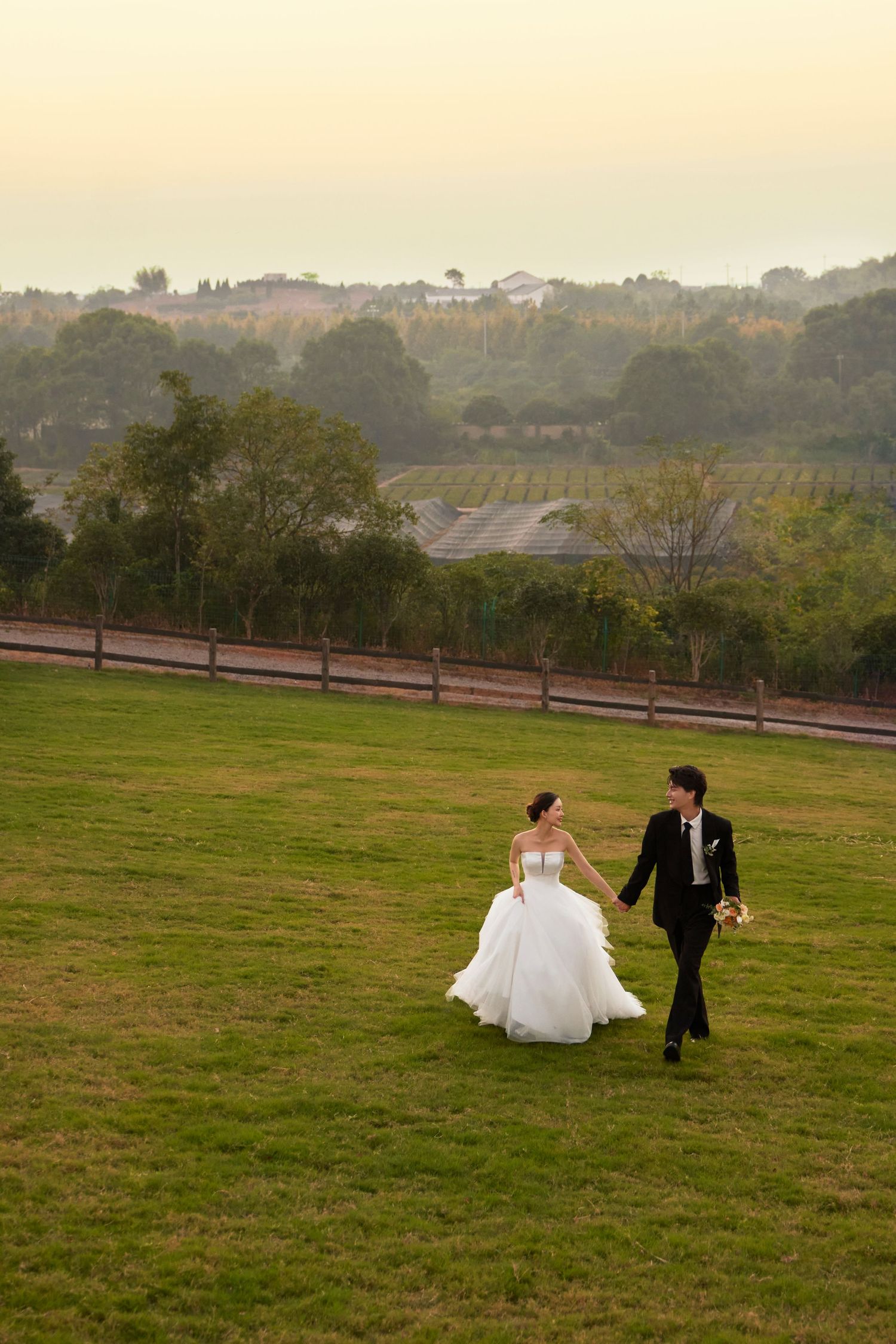 Newlywed couple walks hand-in-hand across green field at dusk with scenic countryside backdrop.