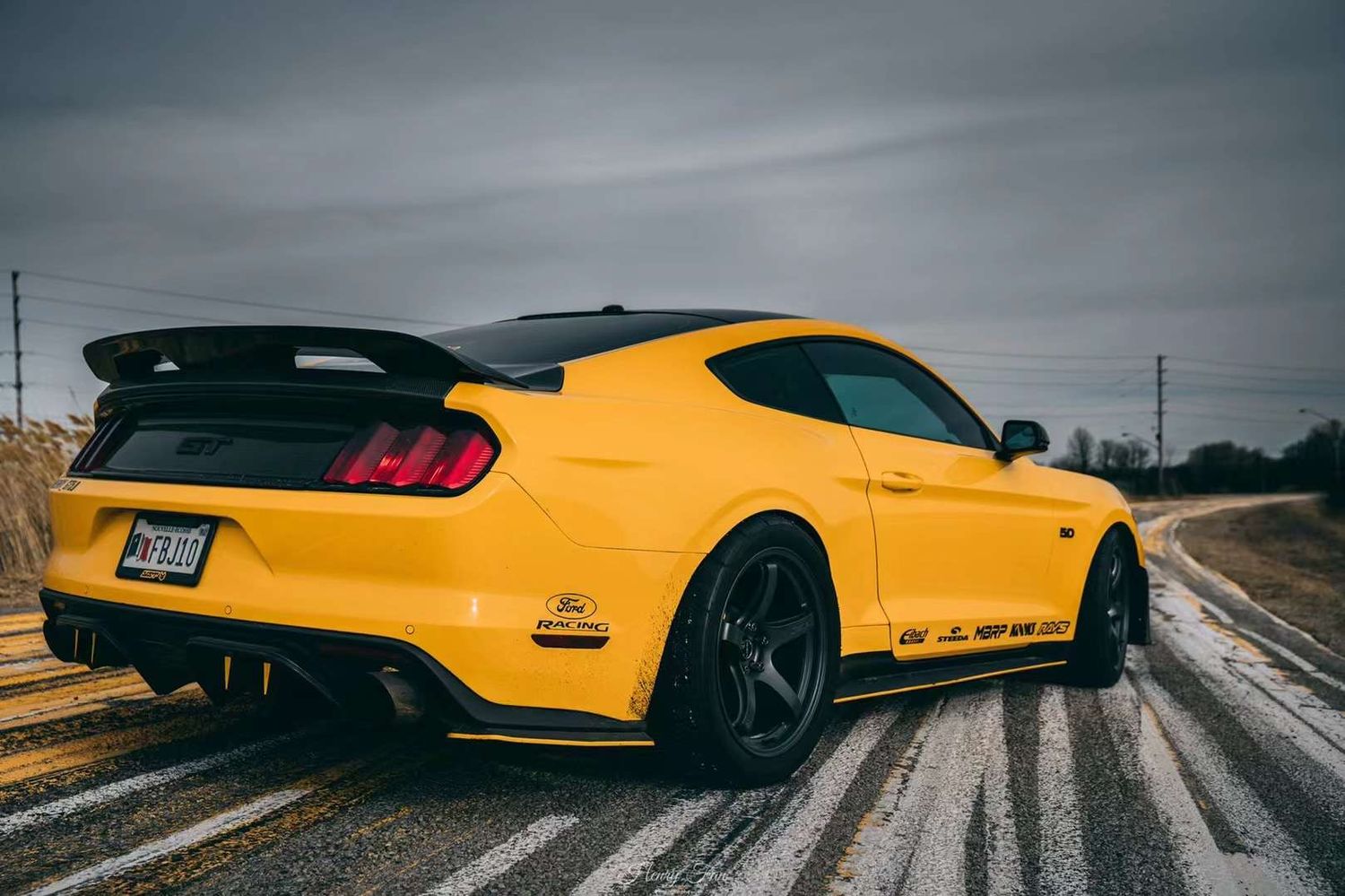 Rear view of a yellow Ford Mustang GT with black spoiler and custom rims on a rural road under overcast skies.
