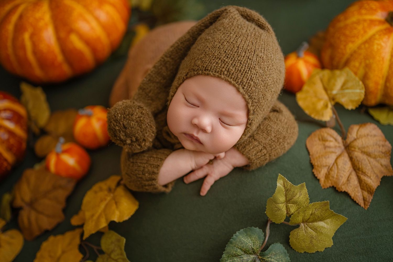 Sleeping newborn wearing brown knit bear bonnet resting on green surface with pumpkins and autumn leaves.