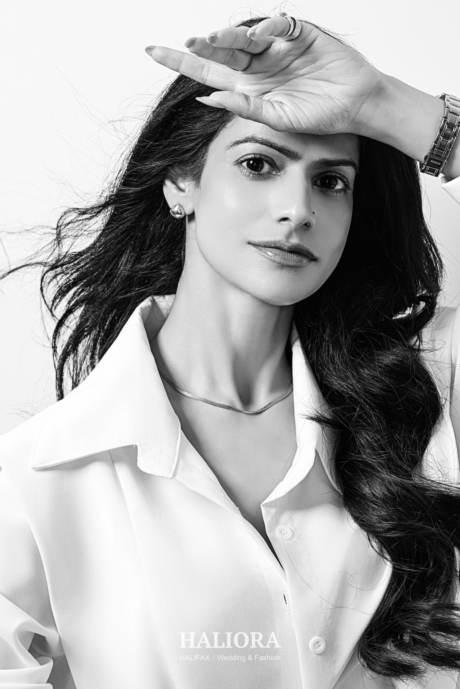 Black and white fashion portrait of woman in white shirt shielding eyes with hand, wearing silver necklace.