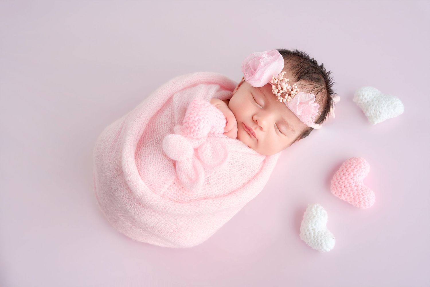Sleeping newborn baby girl wrapped in pink blanket with bow headband surrounded by knitted hearts.