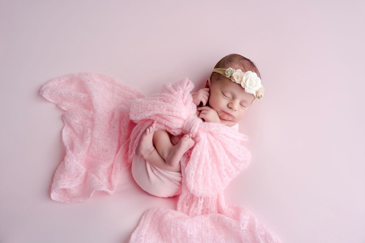 Newborn baby girl sleeping peacefully wrapped in pink lace with a floral headband on a pink background.