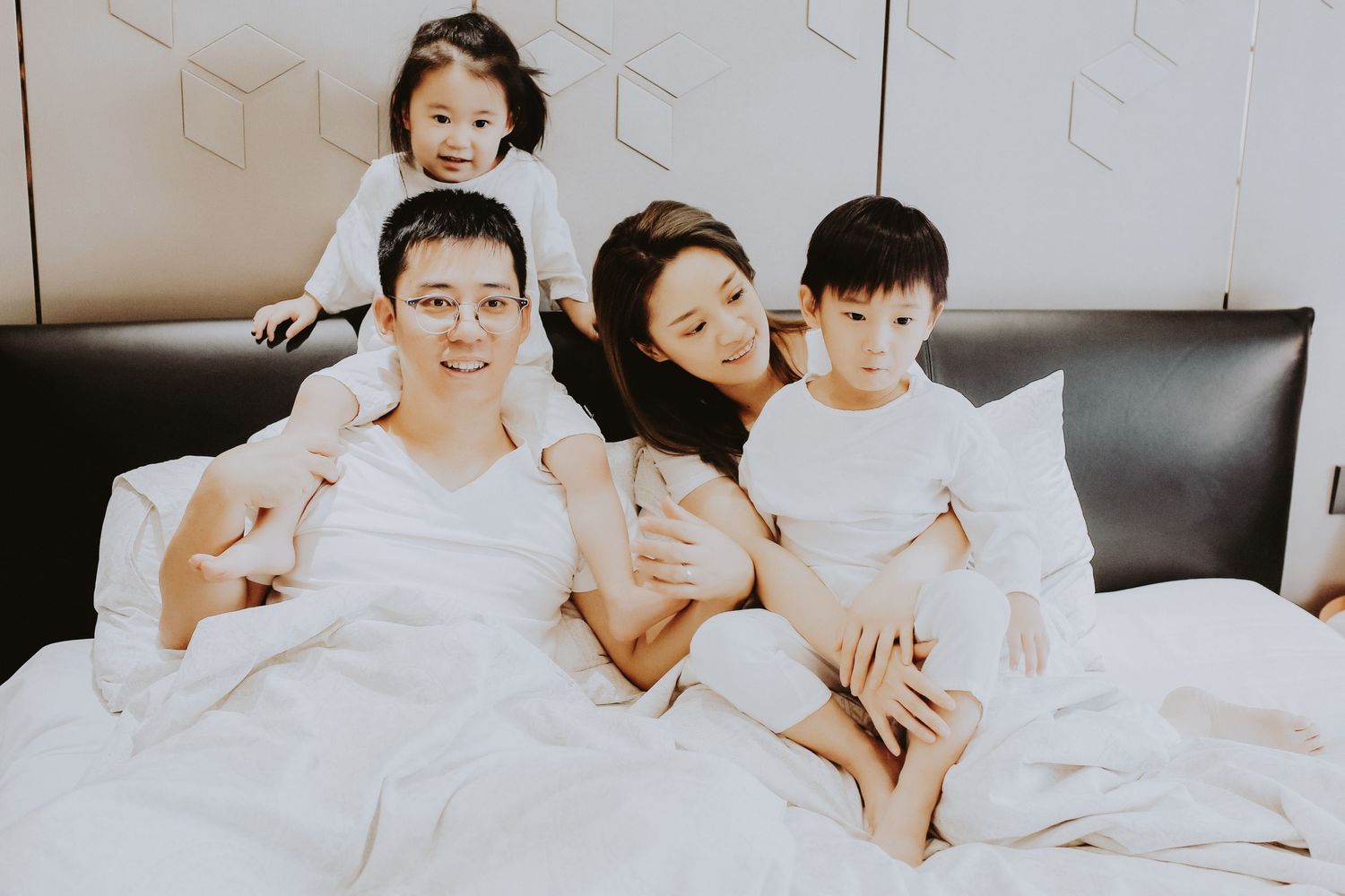 A cheerful Asian family of four relaxes together on a white bed, smiling and enjoying quality time.