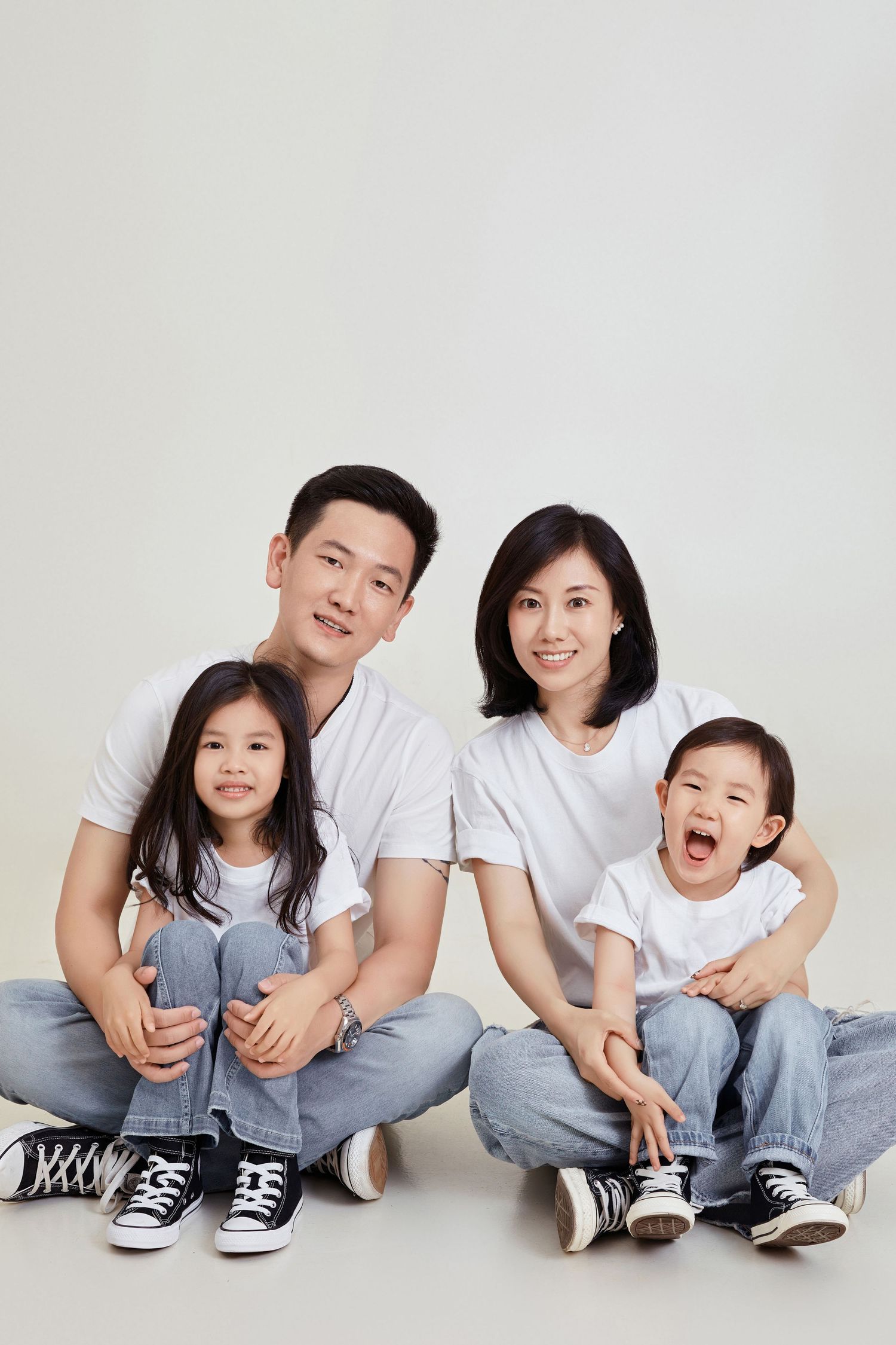 Happy Asian family of four in matching white tees and jeans sitting together smiling in a studio portrait.