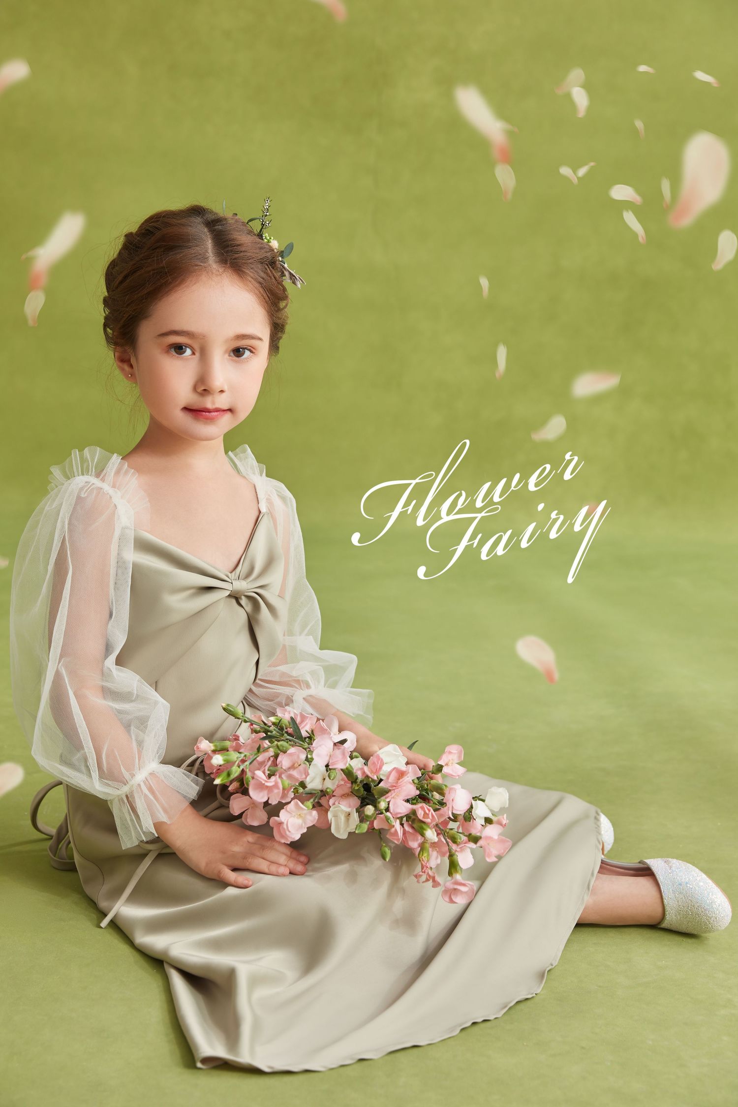 Young girl in sage satin dress with sheer sleeves sits holding pink flowers, petals falling.