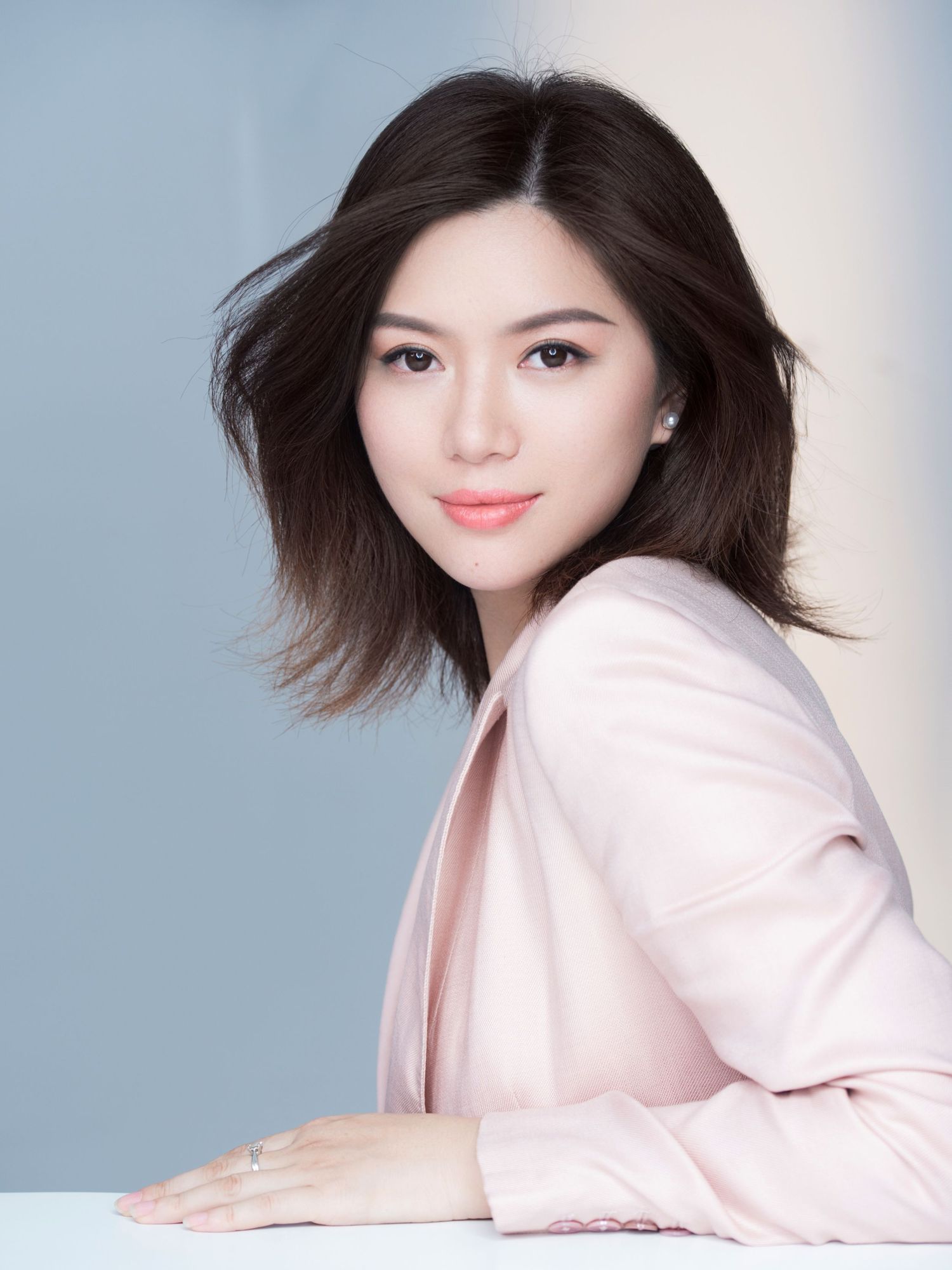 Beautiful Asian woman with windswept dark hair in a pink blazer leaning on a white surface, soft blue backdrop.