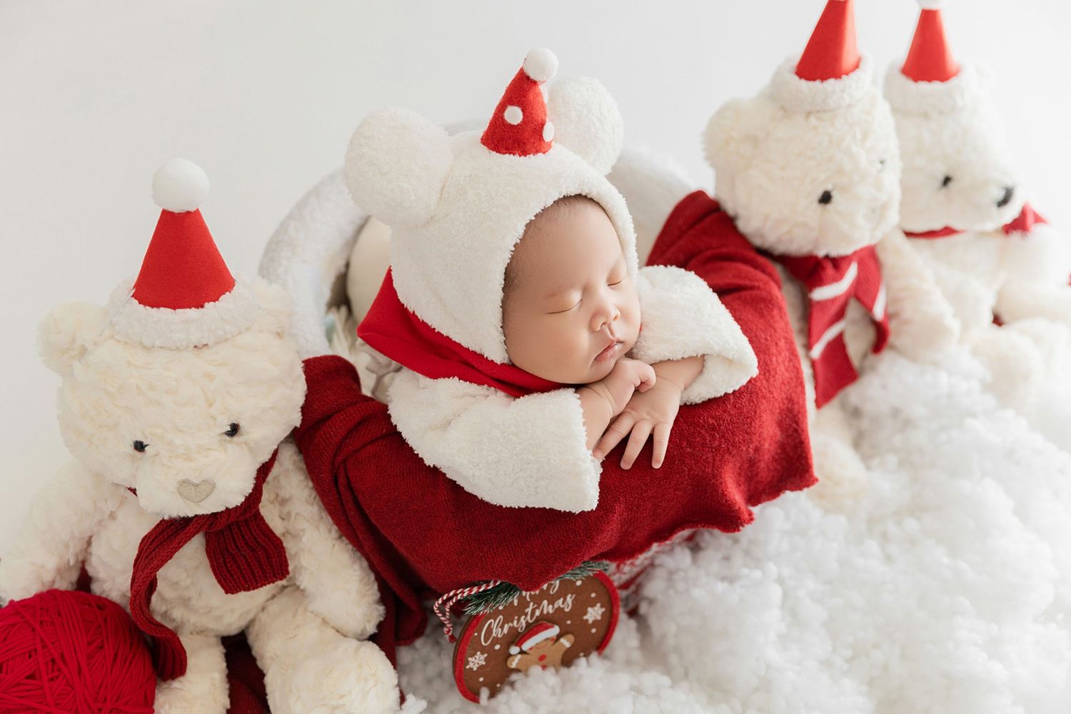 Peaceful sleeping newborn in white Santa costume nestled between festive white teddy bears on snowy white surface.