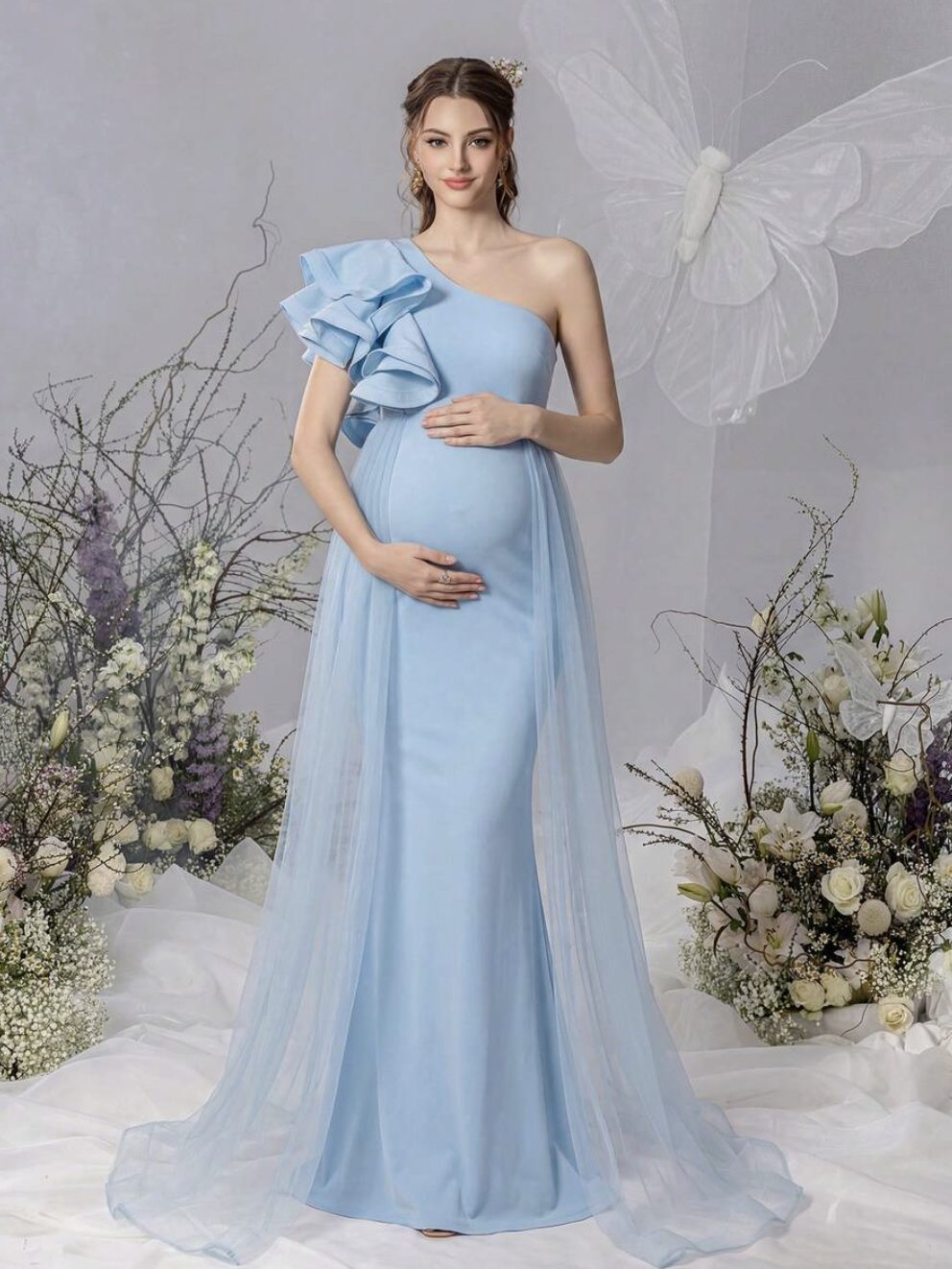 Pregnant woman in elegant light blue one-shoulder ruffle gown with tulle cape train, surrounded by floral decor.