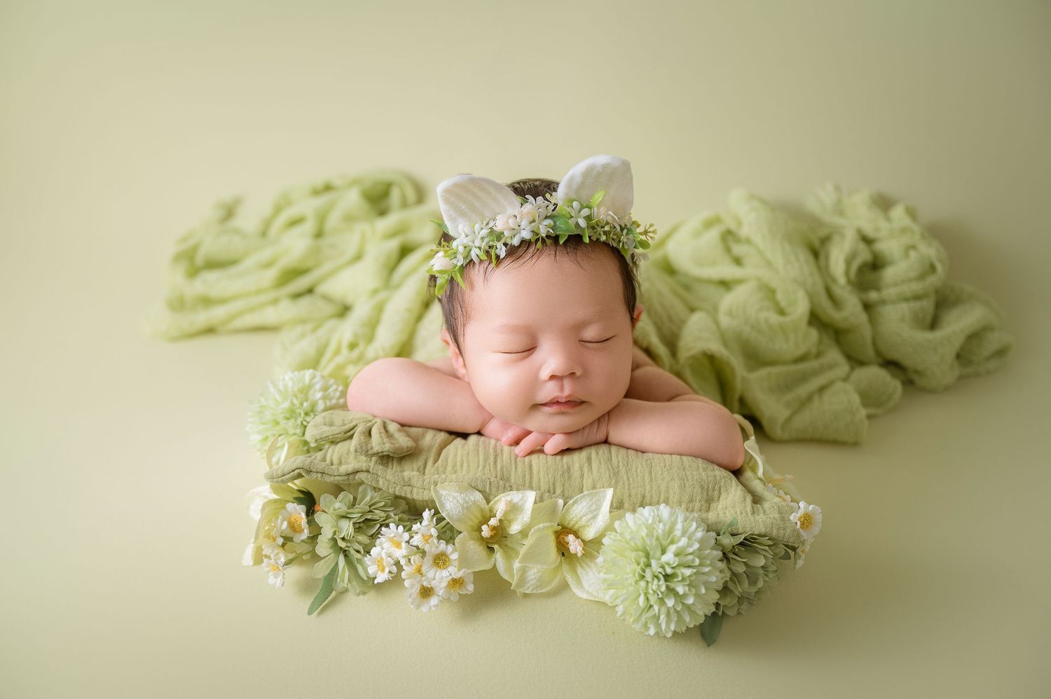 Sleeping newborn baby wearing bunny ear headband on green textured wrap with floral decorations.