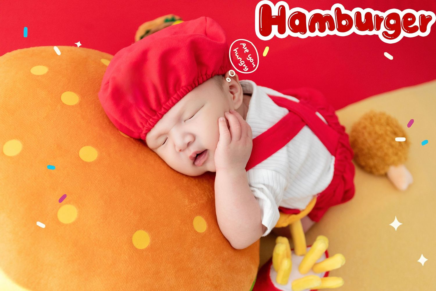 A sleeping baby dressed in a red hamburger costume rests on a giant burger plush toy with fries prop nearby.