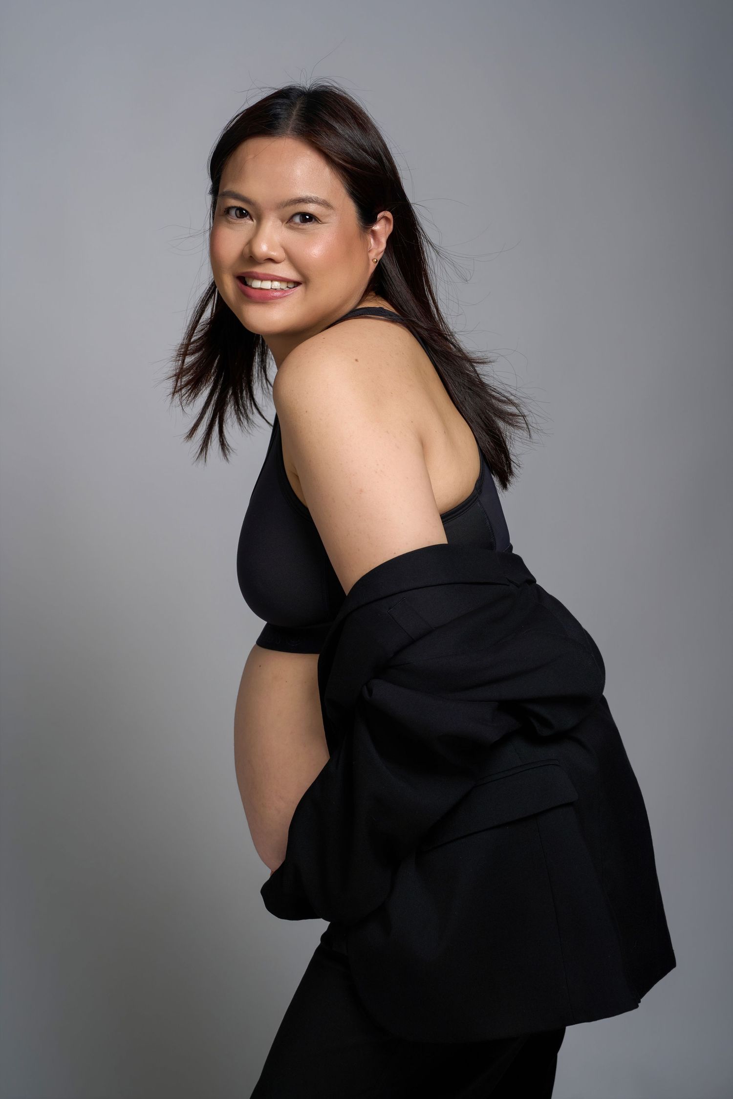 Smiling pregnant woman in black outfit posing sideways, showing baby bump, against gray studio background.