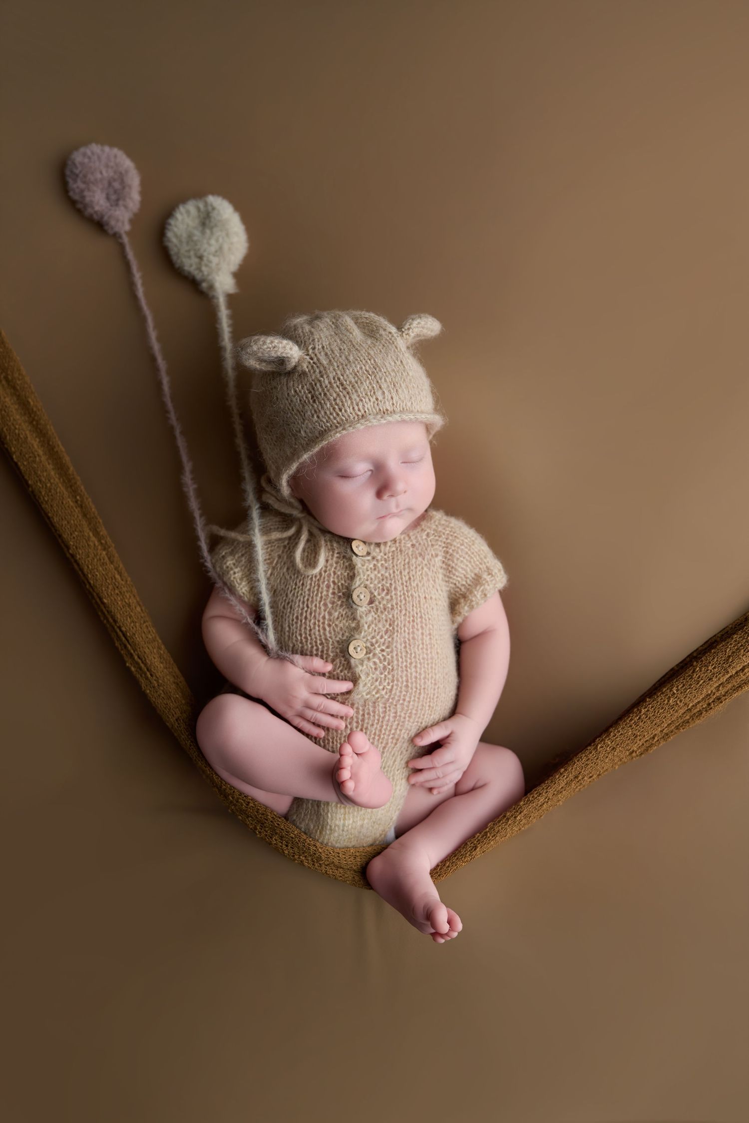 Sleeping newborn baby in beige knit outfit and bear hat with pom-pom balloons, resting in a hammock.