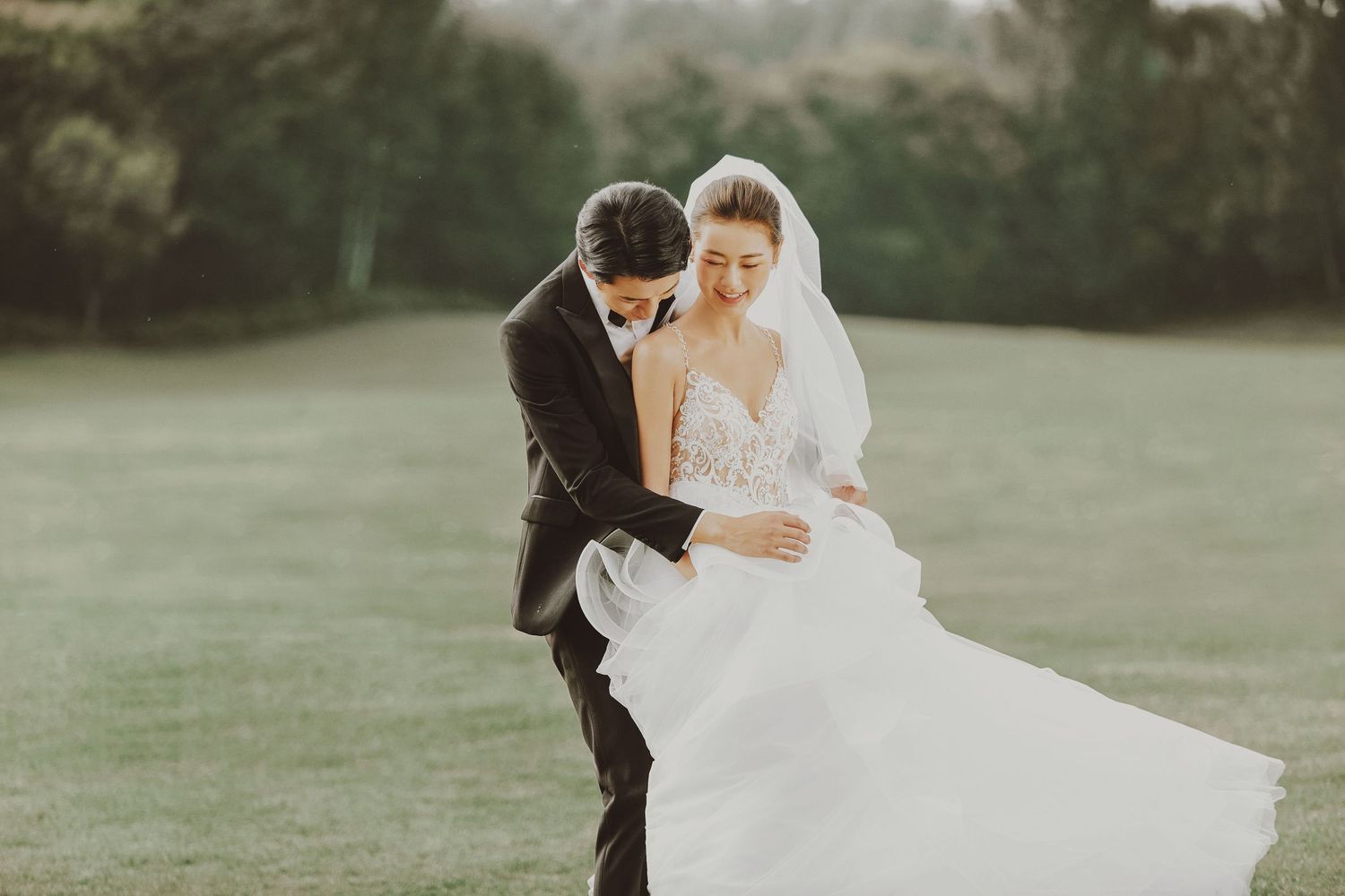 Romantic wedding couple embracing on a lush green lawn, bride in flowing white gown with veil, groom in dark suit.