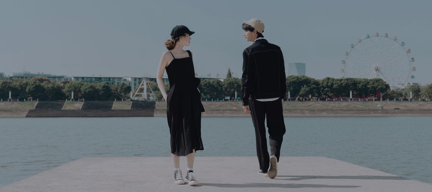 A stylish couple in black outfits walking together on a waterfront pier with a Ferris wheel in the background.