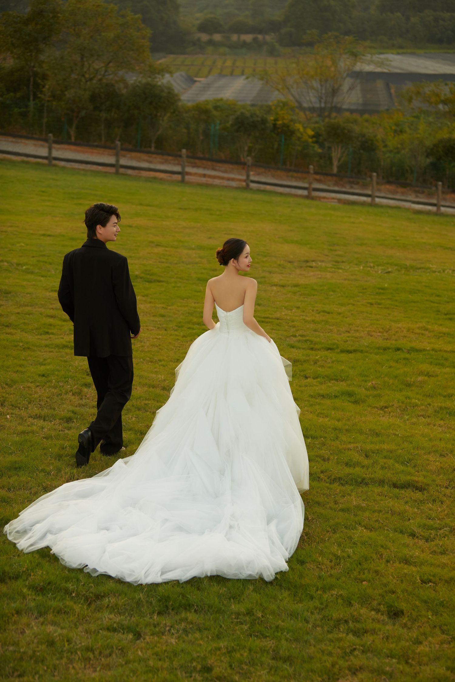 Bride in flowing white gown with long train walks with groom in black suit across green field.