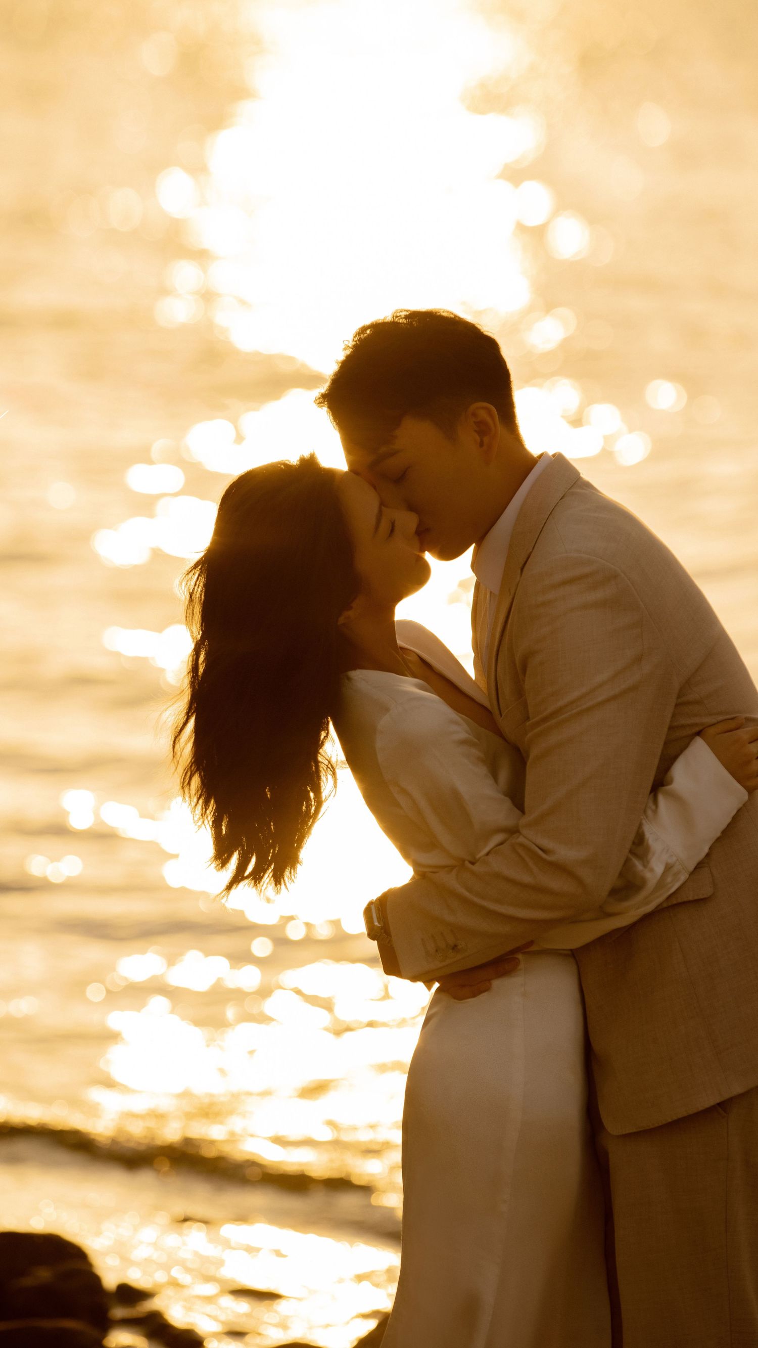 Romantic couple sharing a kiss at golden sunset by the ocean in elegant wedding attire.