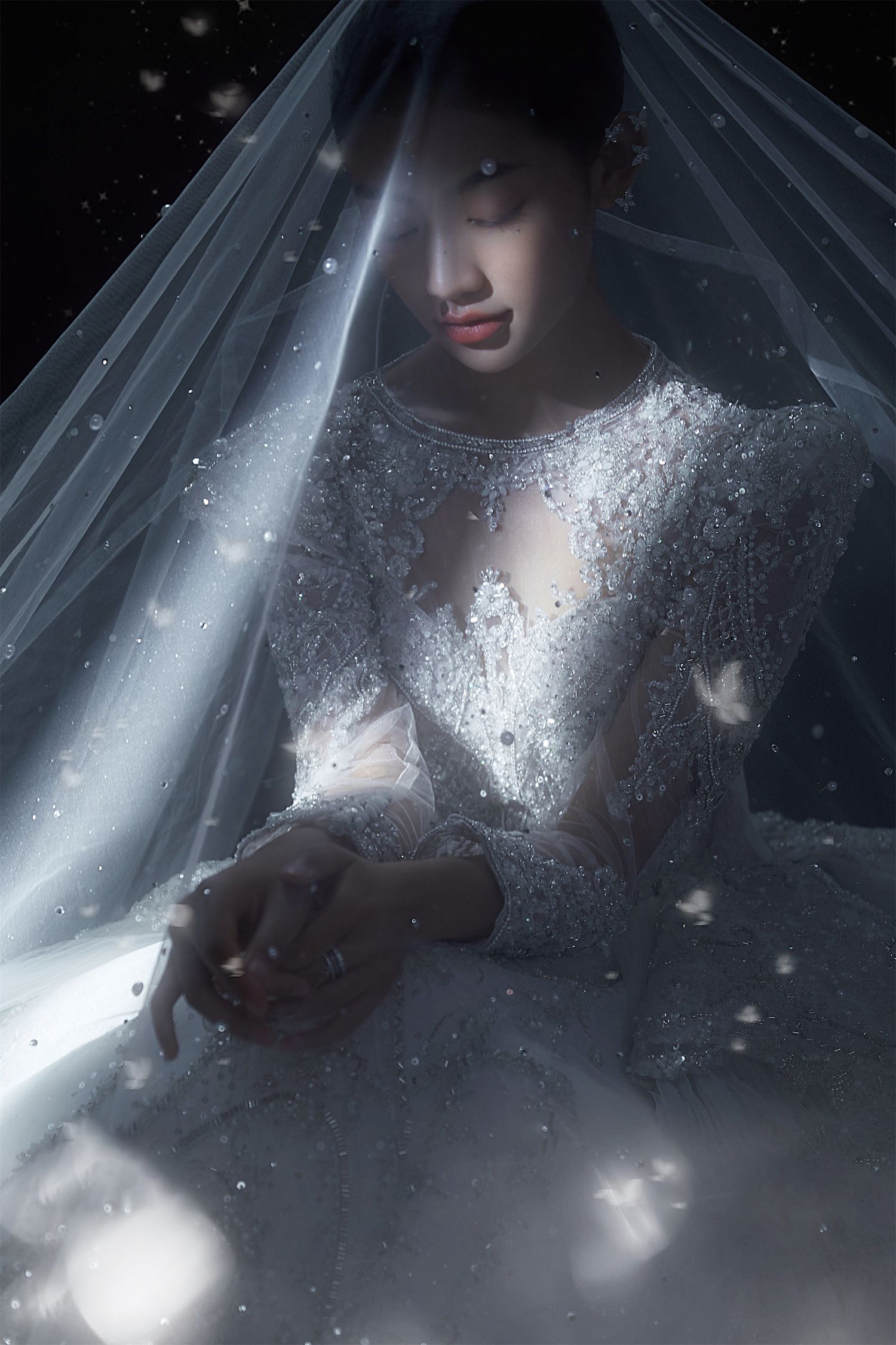 Bride in beaded white wedding gown under sheer veil with sparkling light bokeh on dark background.