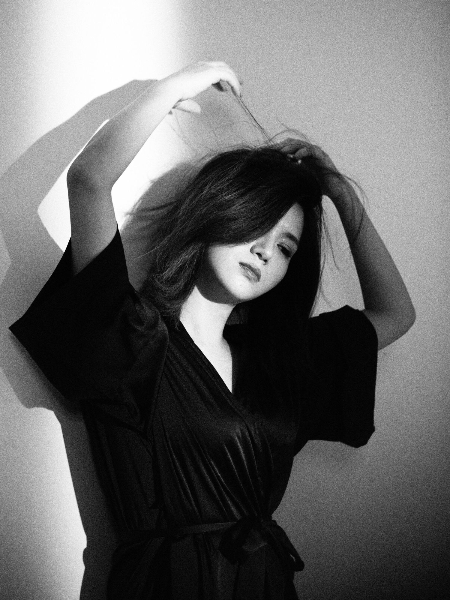 Black and white artistic photo of an Asian woman in a dark wrap robe with arms raised above her head.