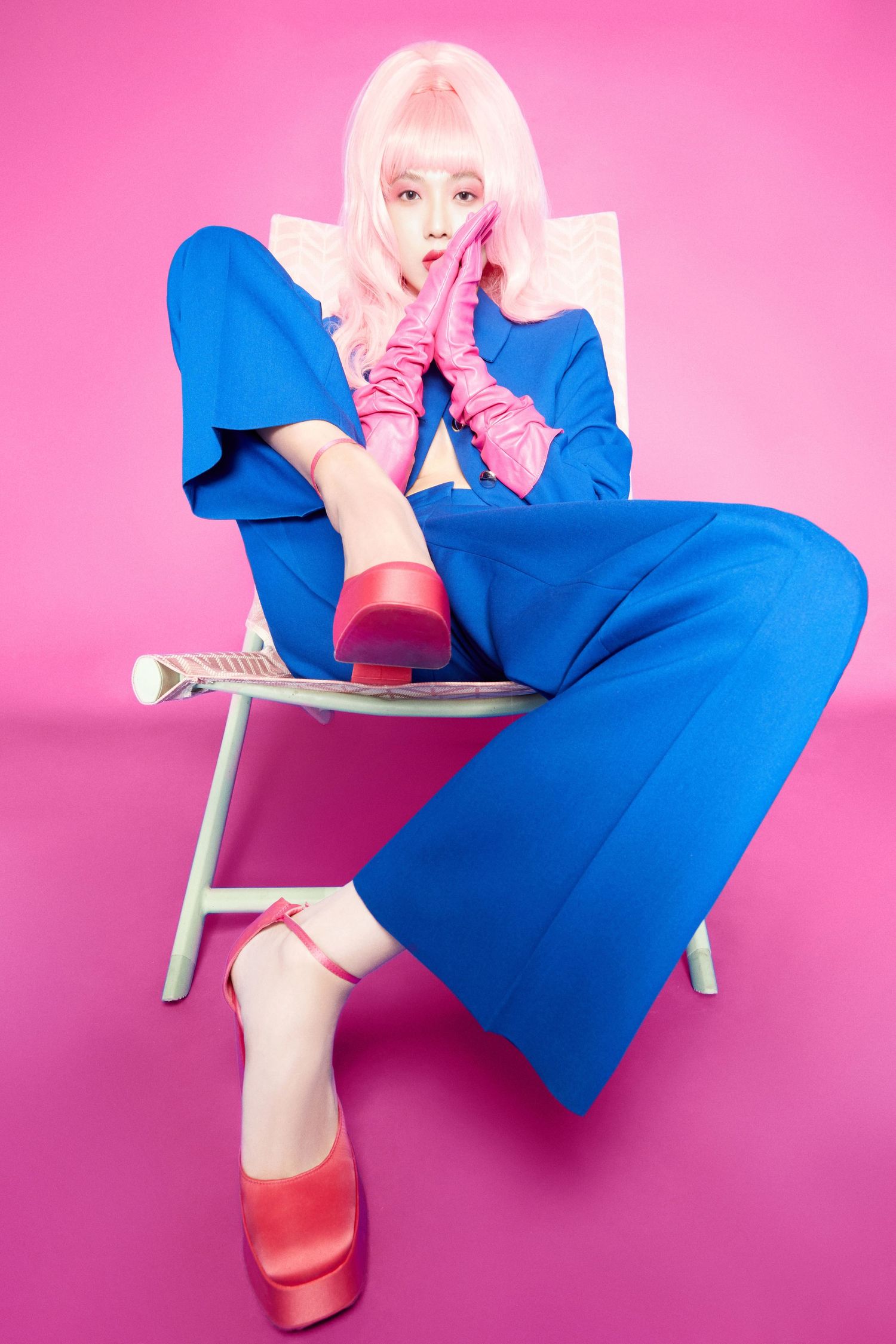 Model in blue wide-leg suit, pink wig, gloves, and platform heels poses on chair against pink background.