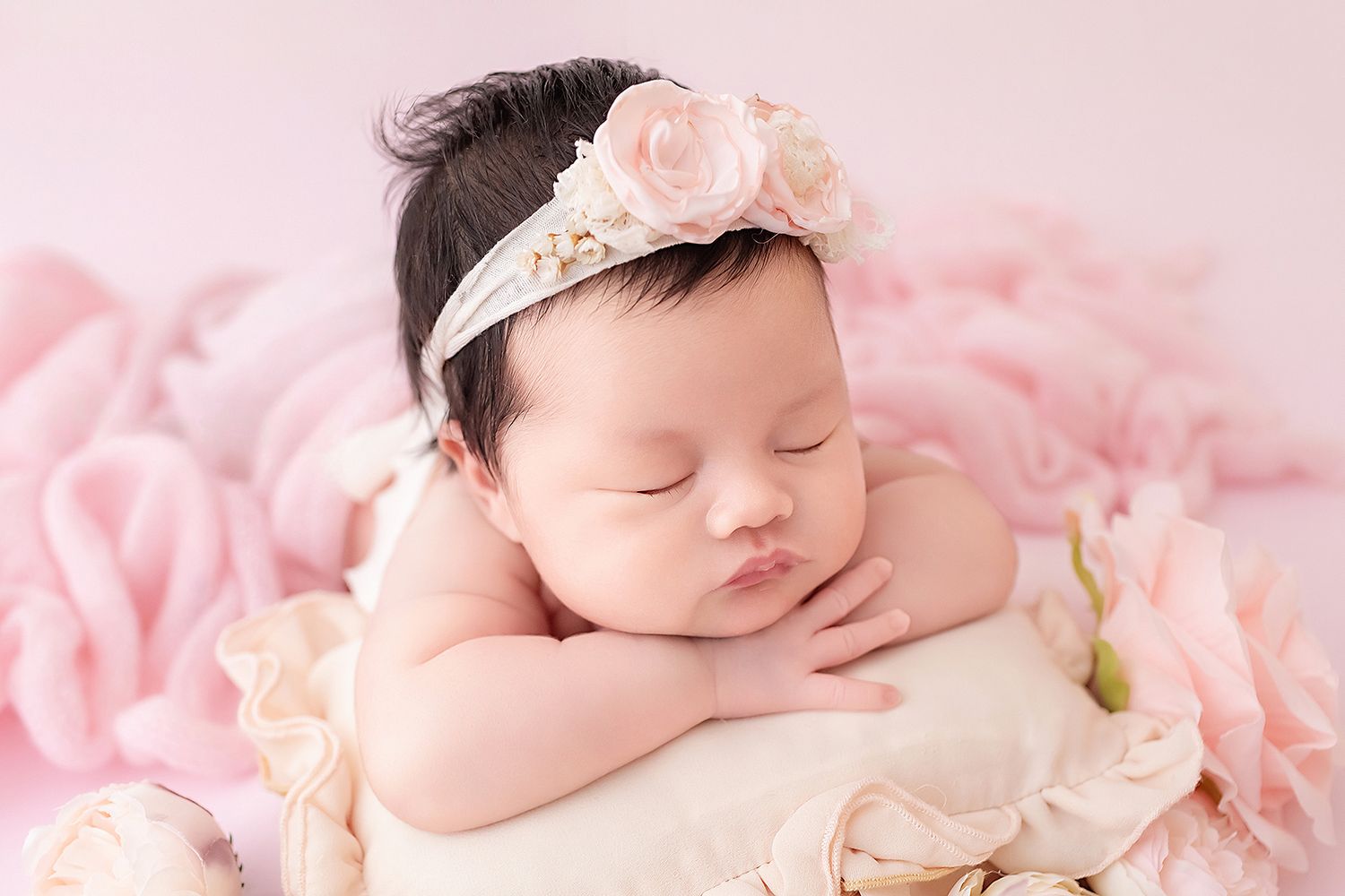 Peaceful newborn baby girl resting chin on hands wearing pink floral headband among roses.