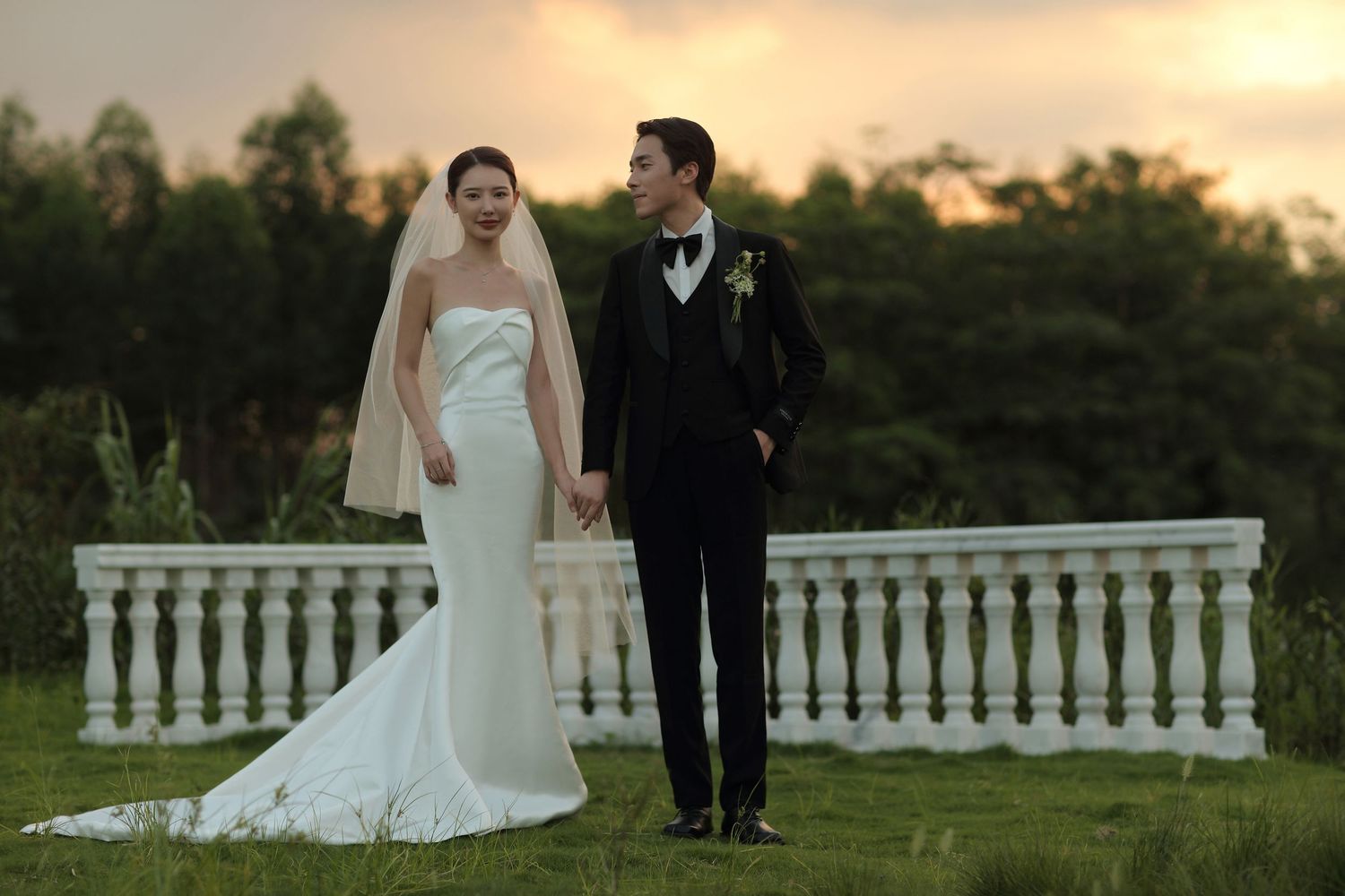Newlyweds in classic black and white wedding attire hold hands on green lawn at golden hour.