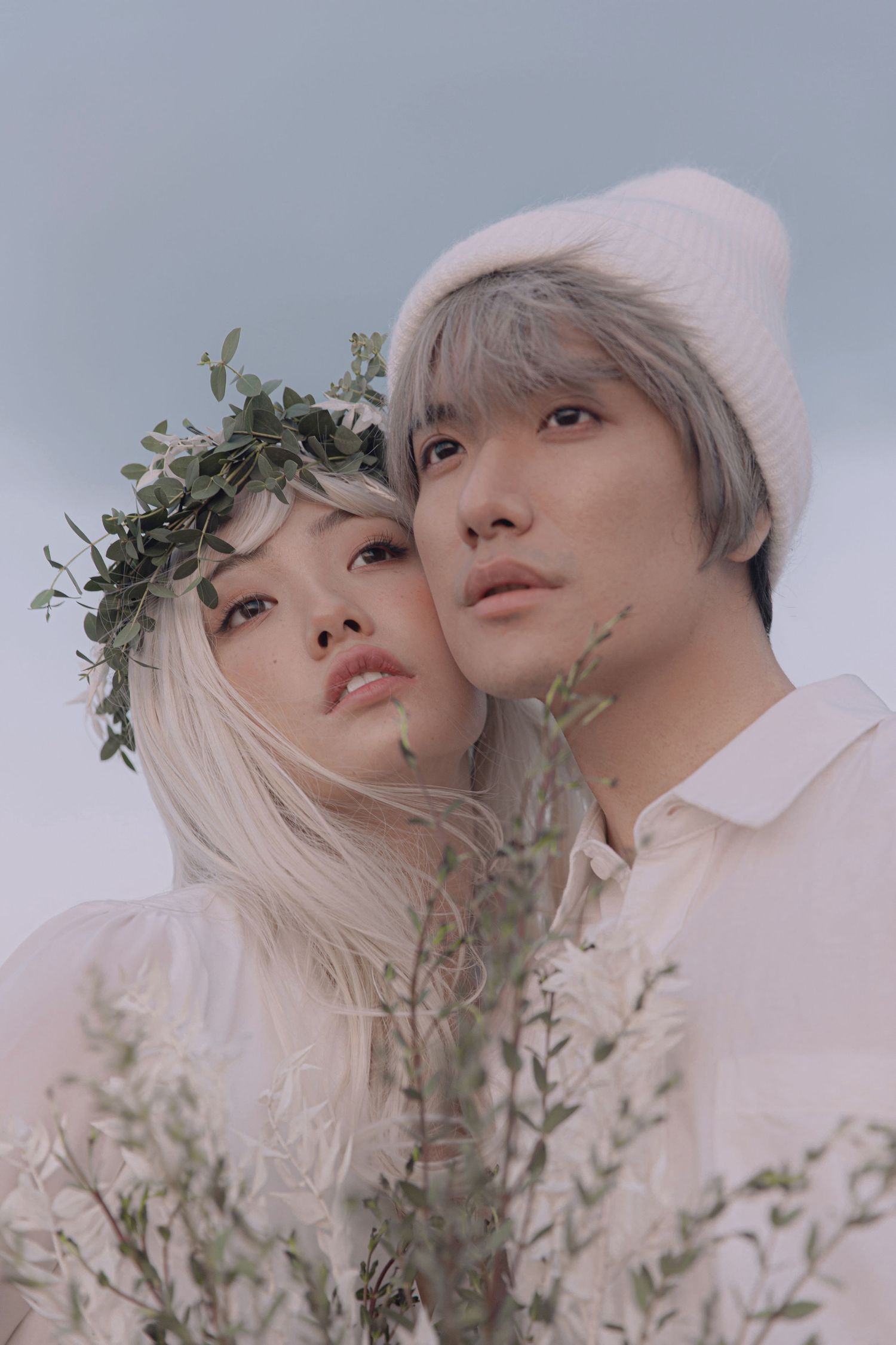Young couple in white clothing with woman wearing leaf crown leans against man in white beanie outdoors.