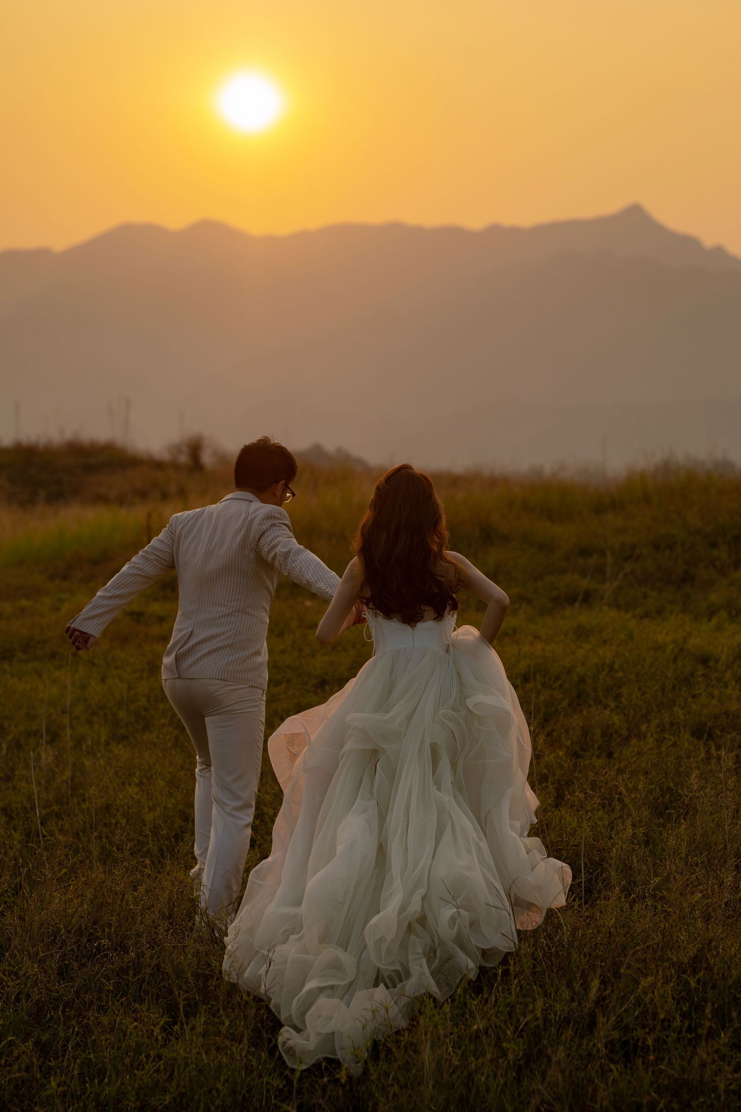 Bride and groom walk away hand in hand into a glowing golden sunset over mountain hills in wedding attire.