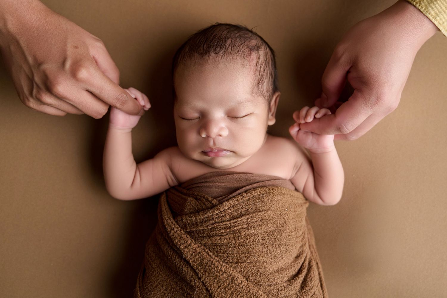 Newborn baby swaddled in brown wrap sleeping peacefully while parents gently hold their tiny hands.