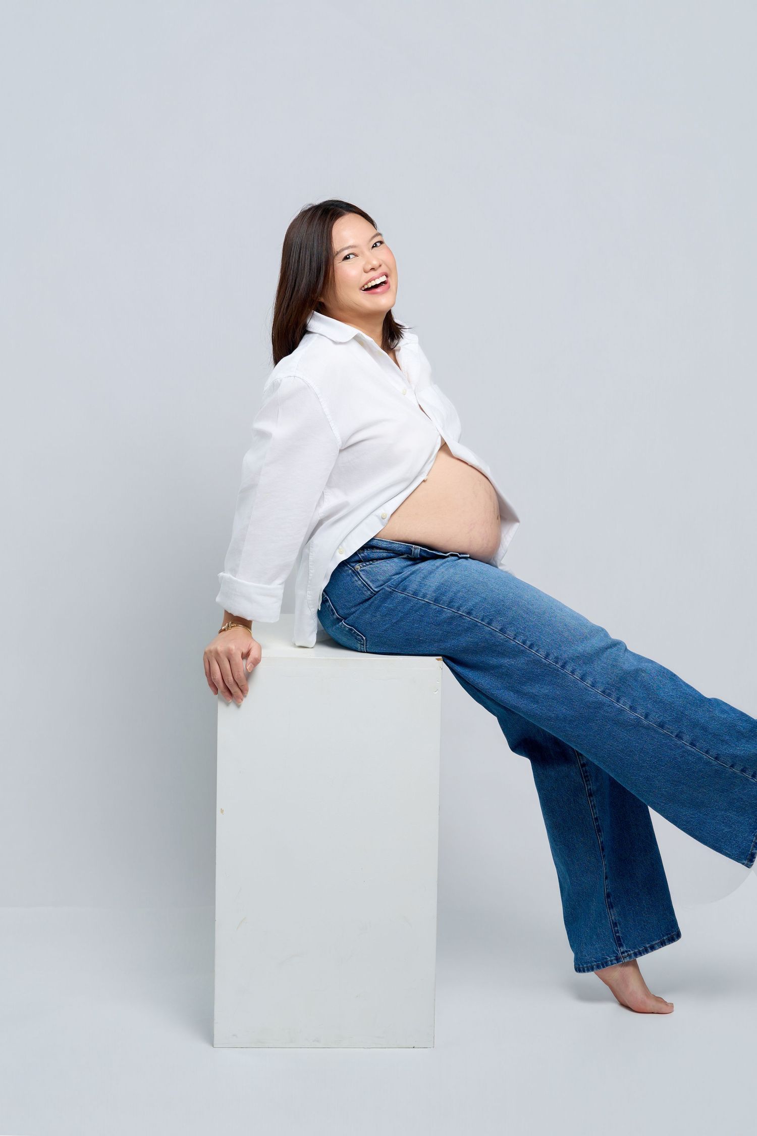 Smiling pregnant woman in white shirt and jeans sitting on white cube, showing baby bump against gray background.