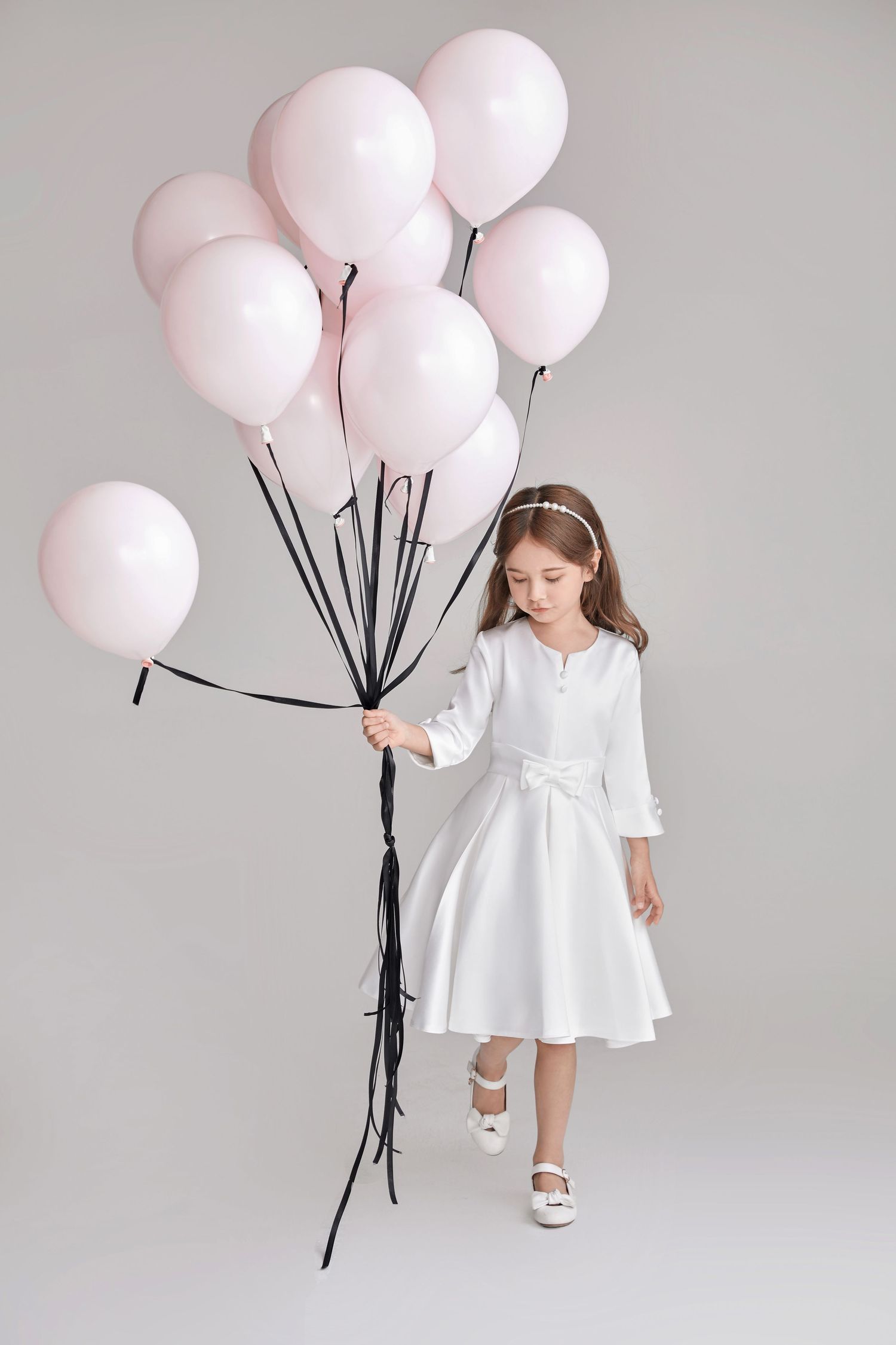 Young girl in white dress holding pink balloons on gray background.