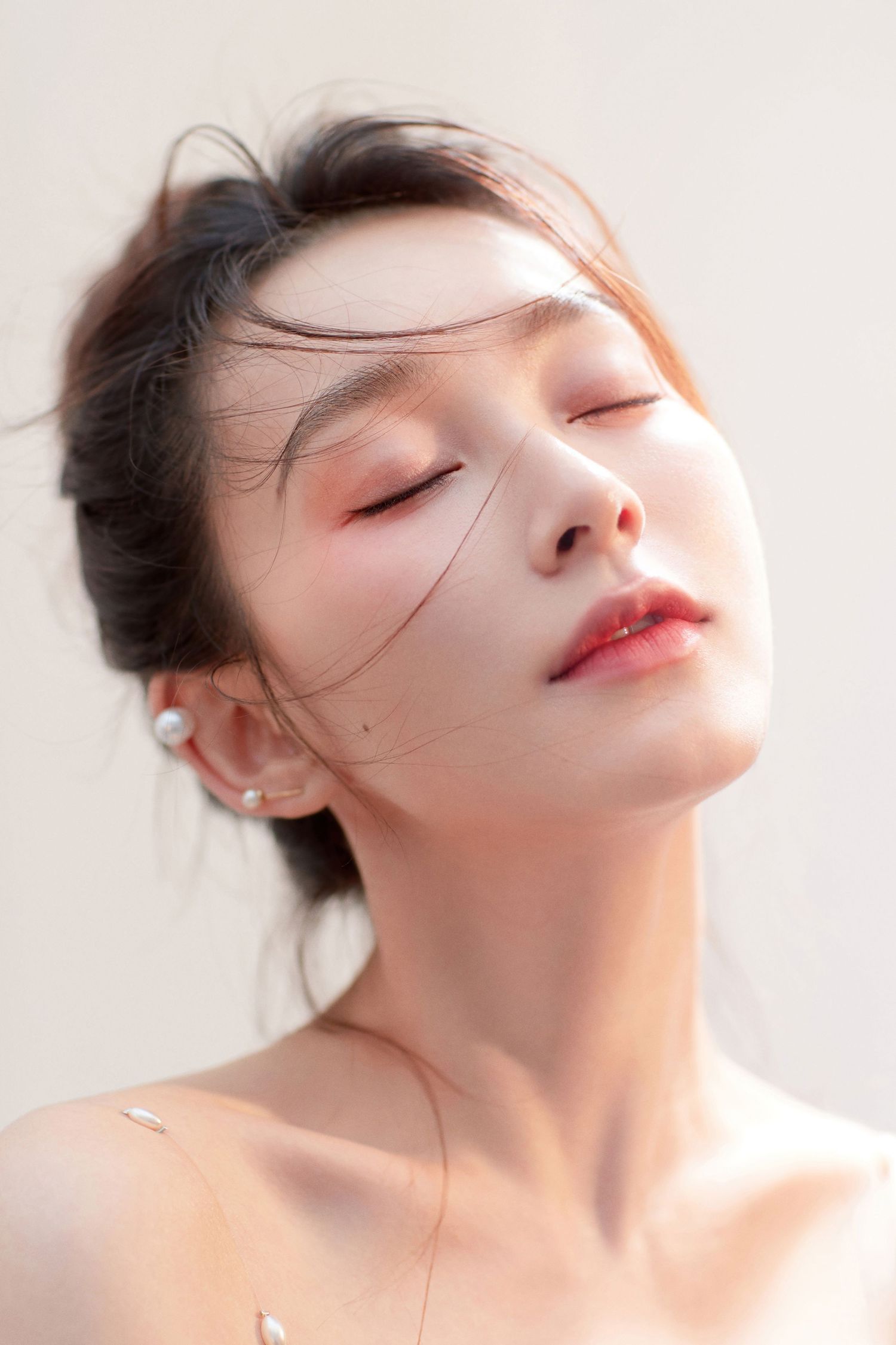 Close-up portrait of an Asian woman with closed eyes, pearl earrings, and bare shoulders in soft natural light.