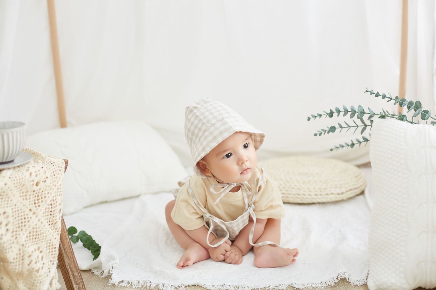 An adorable baby wearing a checkered bonnet sits on a white blanket in a softly lit, cozy room.