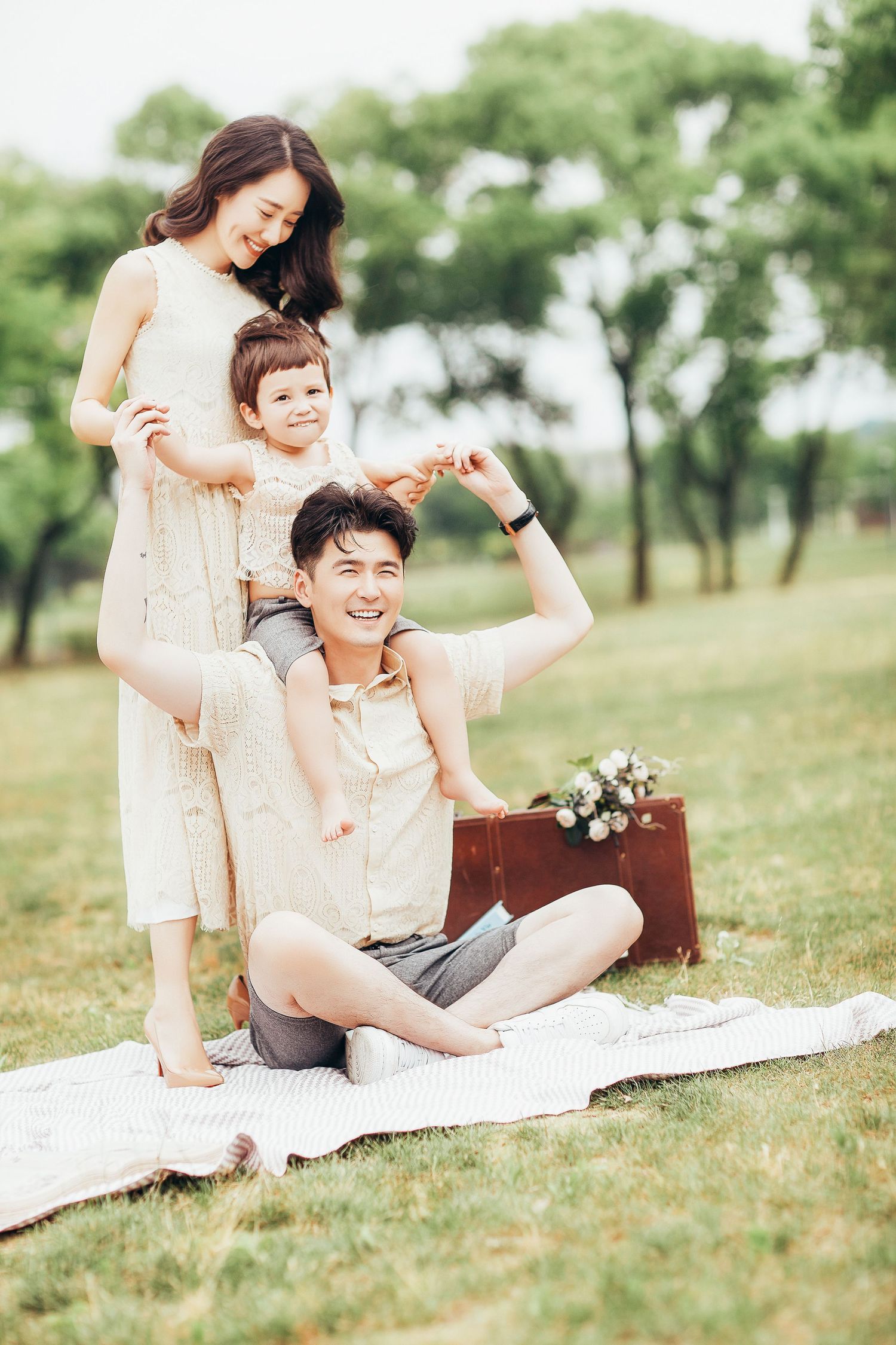 A cheerful Asian family poses on a picnic blanket as a young boy sits on his fathers shoulders.