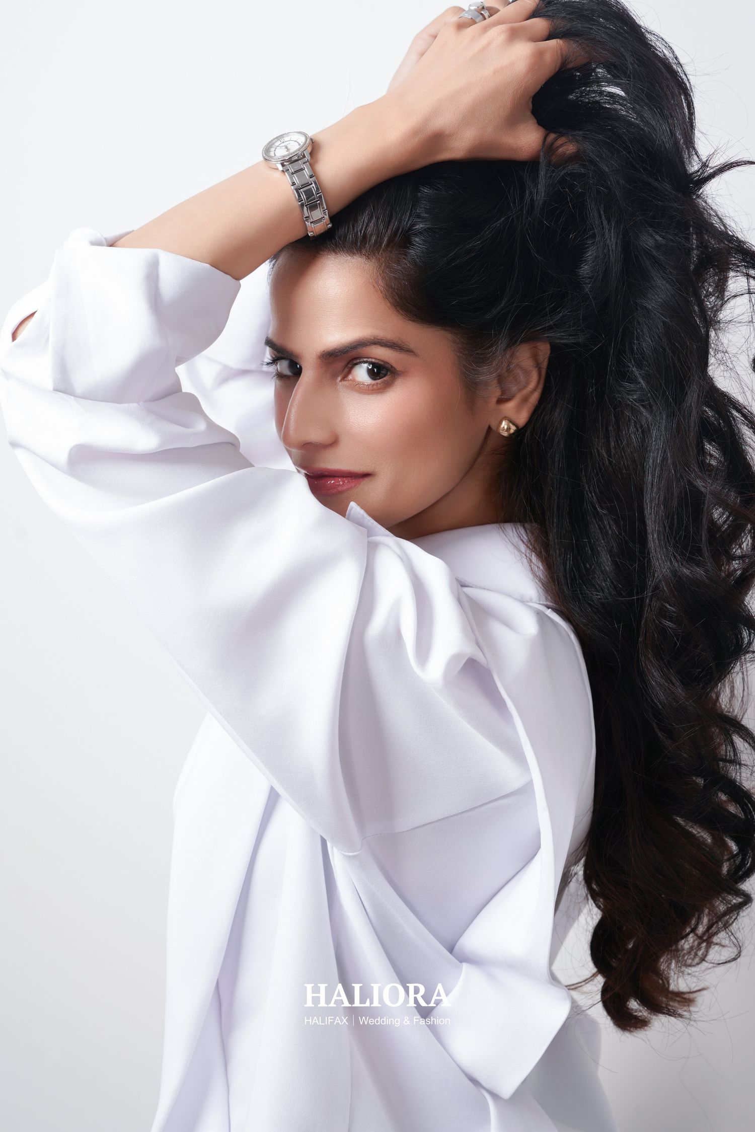 Young woman in white oversized shirt holding her dark wavy hair up, wearing a silver watch, fashion editorial.
