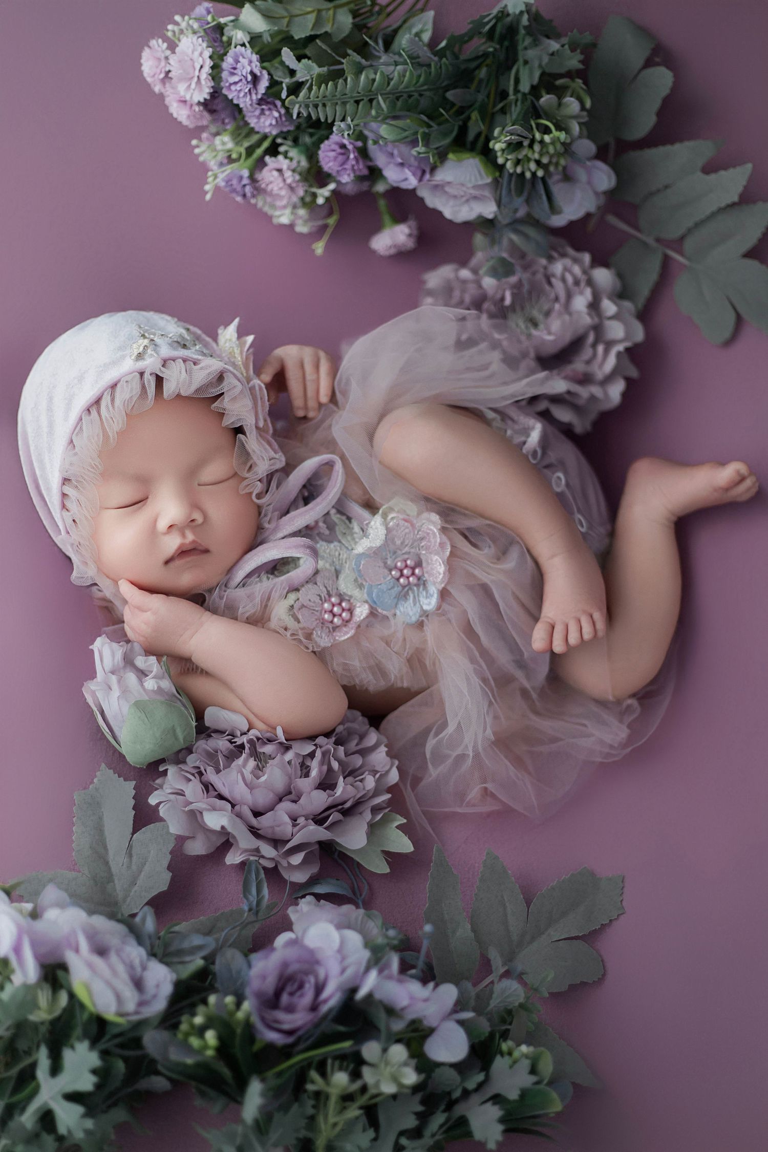 Peaceful newborn baby girl in lilac dress and bonnet posed among purple flowers and green leaves on mauve backdrop.