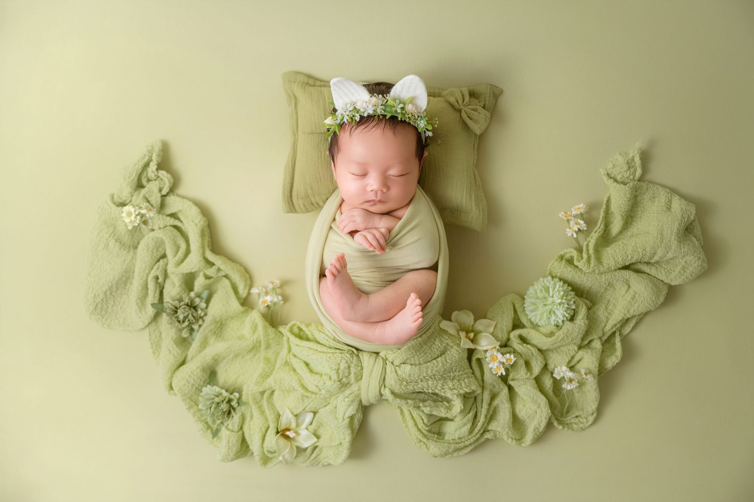 Newborn baby swaddled in green wrap sleeping peacefully on green blanket with floral bunny ear headband.