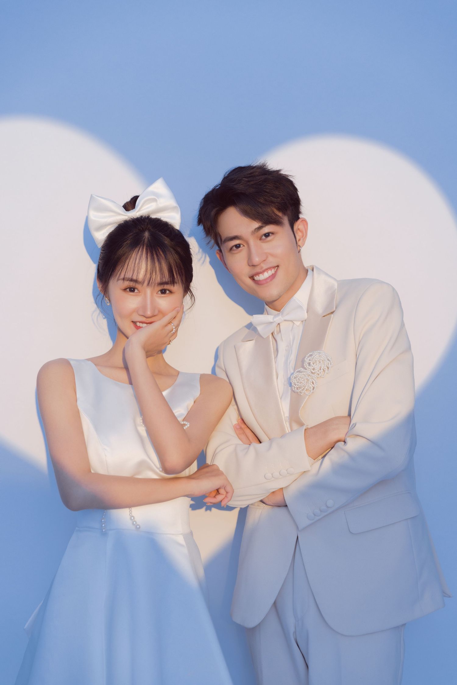 A cheerful couple in white wedding outfits pose together against a soft blue background with heart shadows.