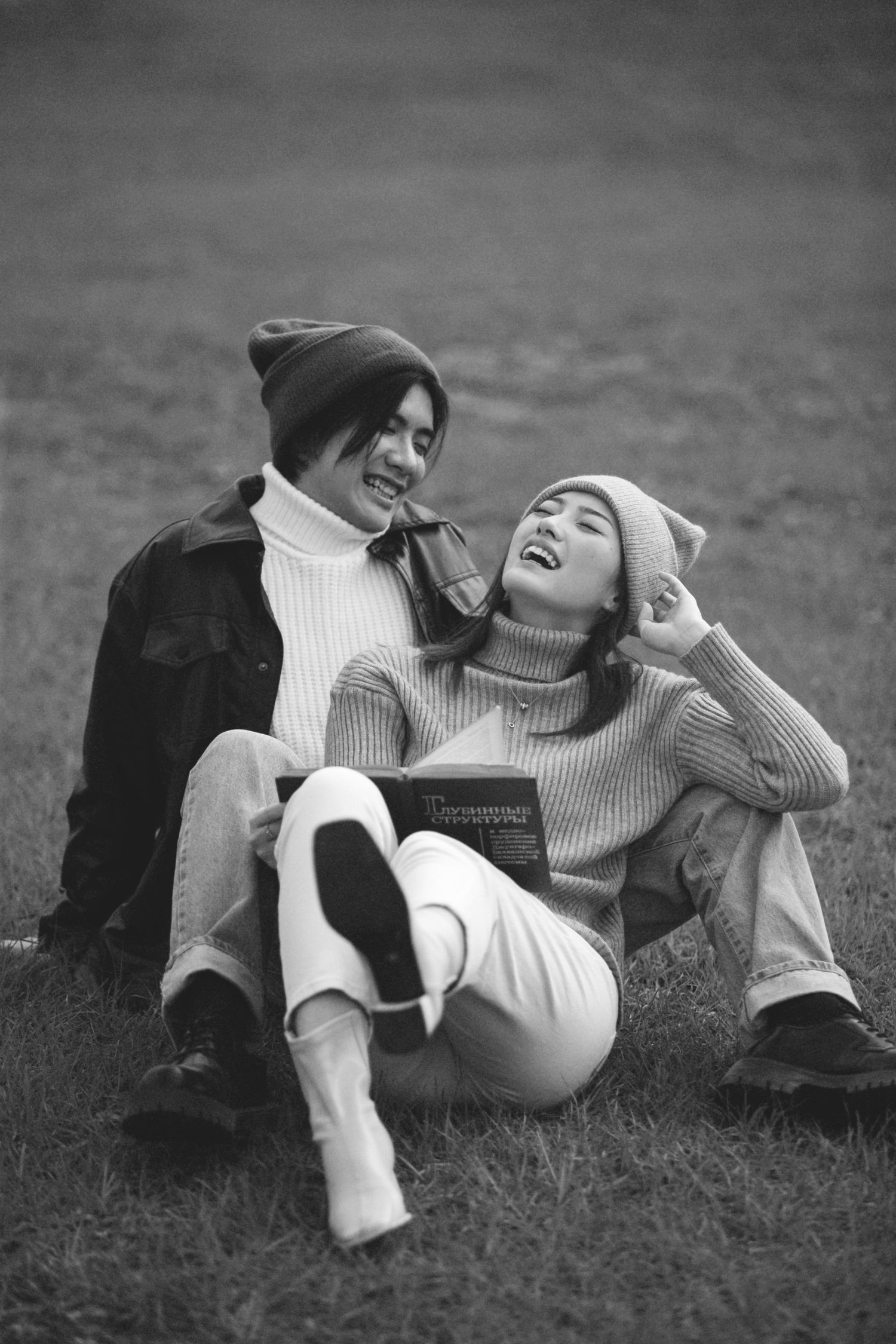 Happy couple sitting on grass laughing together, wearing cozy sweaters and beanies in black and white photo.