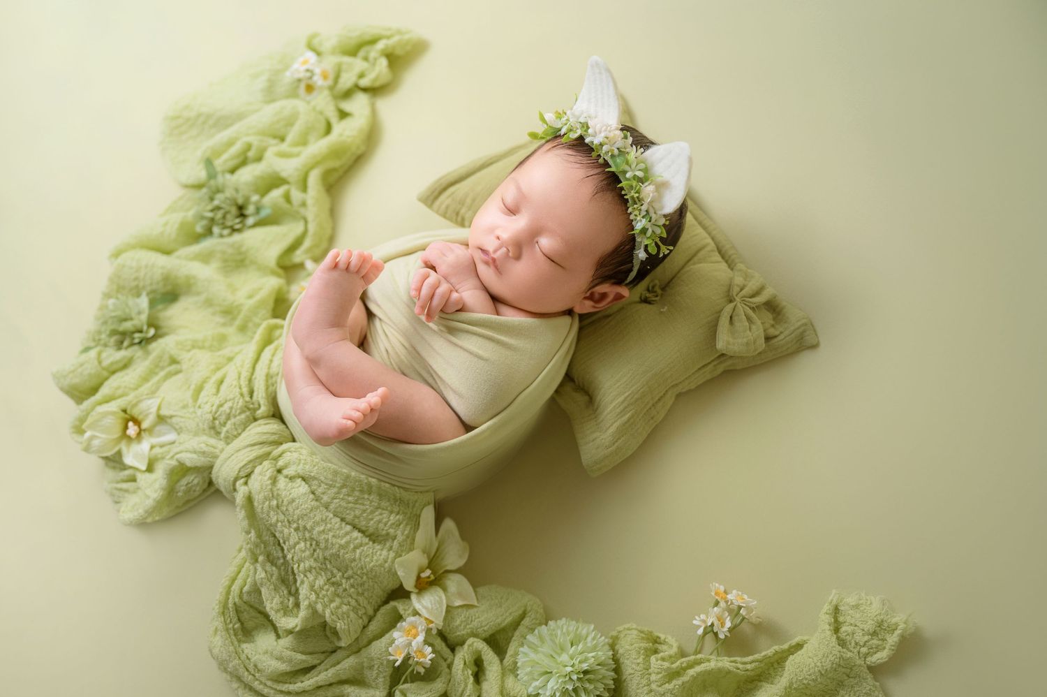 Newborn baby sleeping peacefully in green wrap with floral headband on green background.
