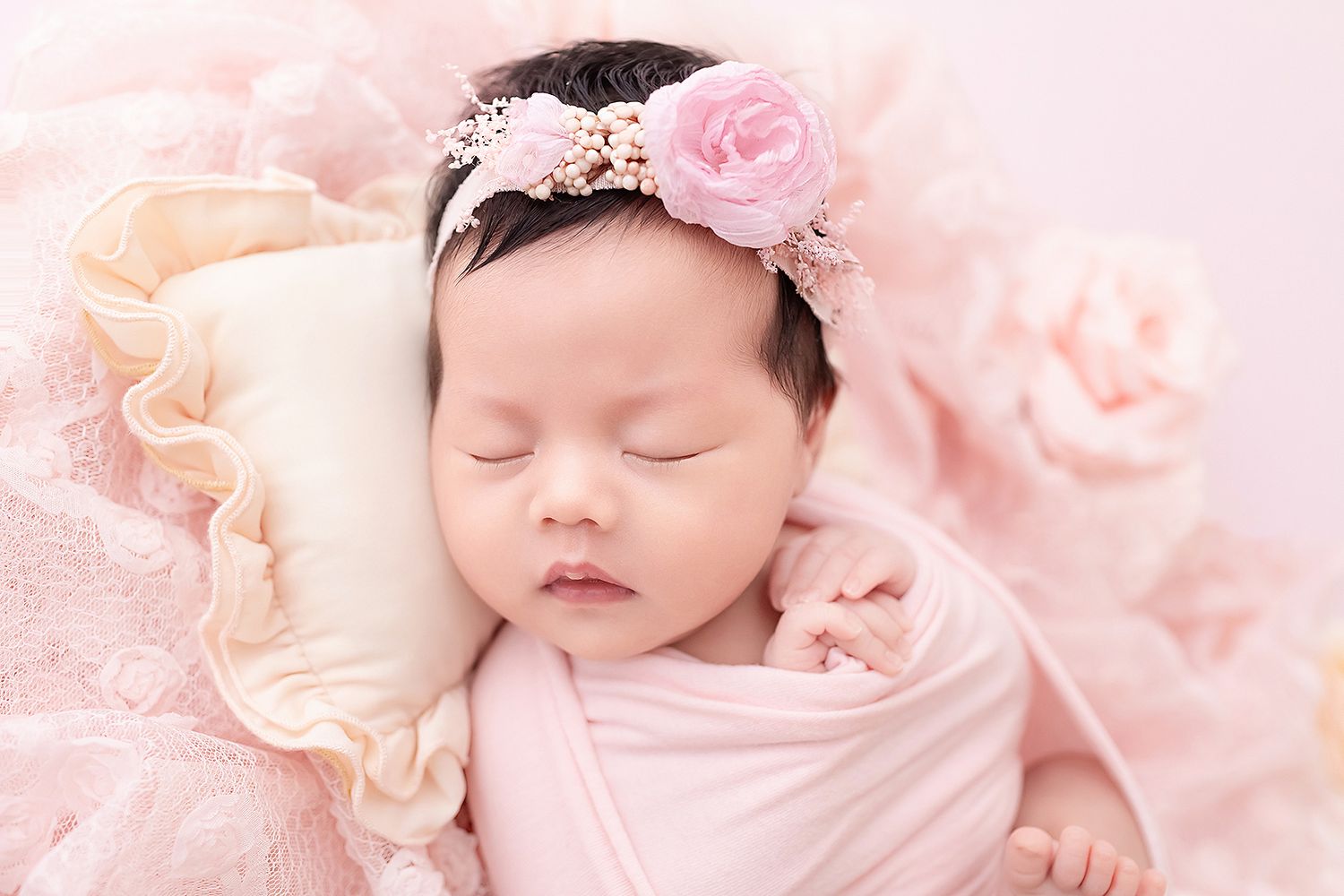 Sleeping newborn baby girl swaddled in pink wrap wearing floral rose headband on pink backdrop.
