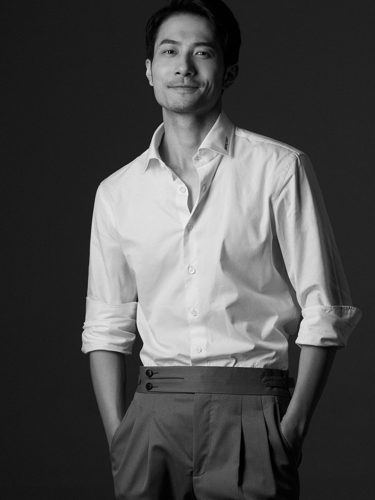 Stylish Asian man in white button-up shirt and grey trousers posing confidently in black and white portrait.