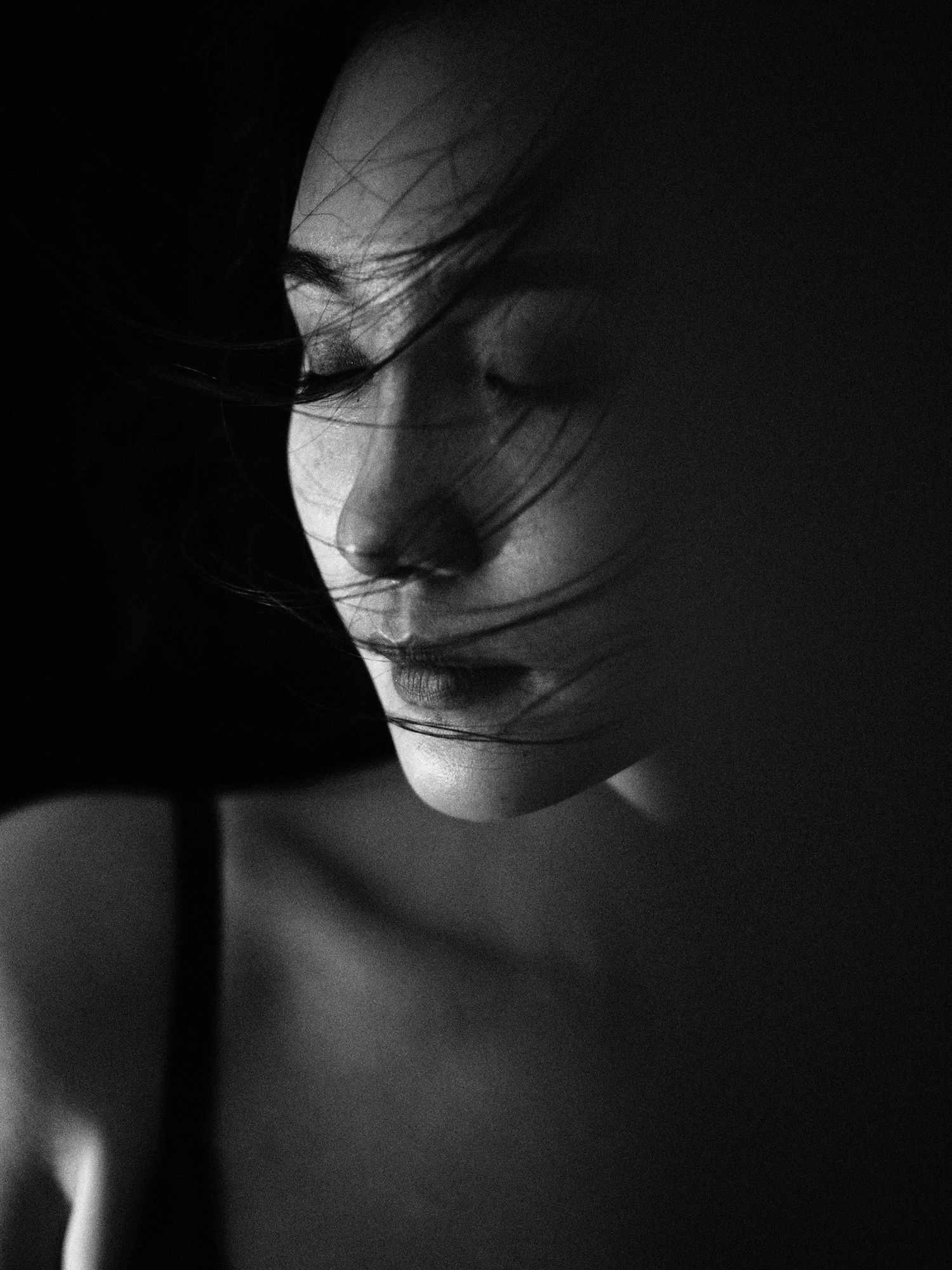Dramatic black and white close-up of a woman with eyes closed and hair blowing across her face in moody lighting.