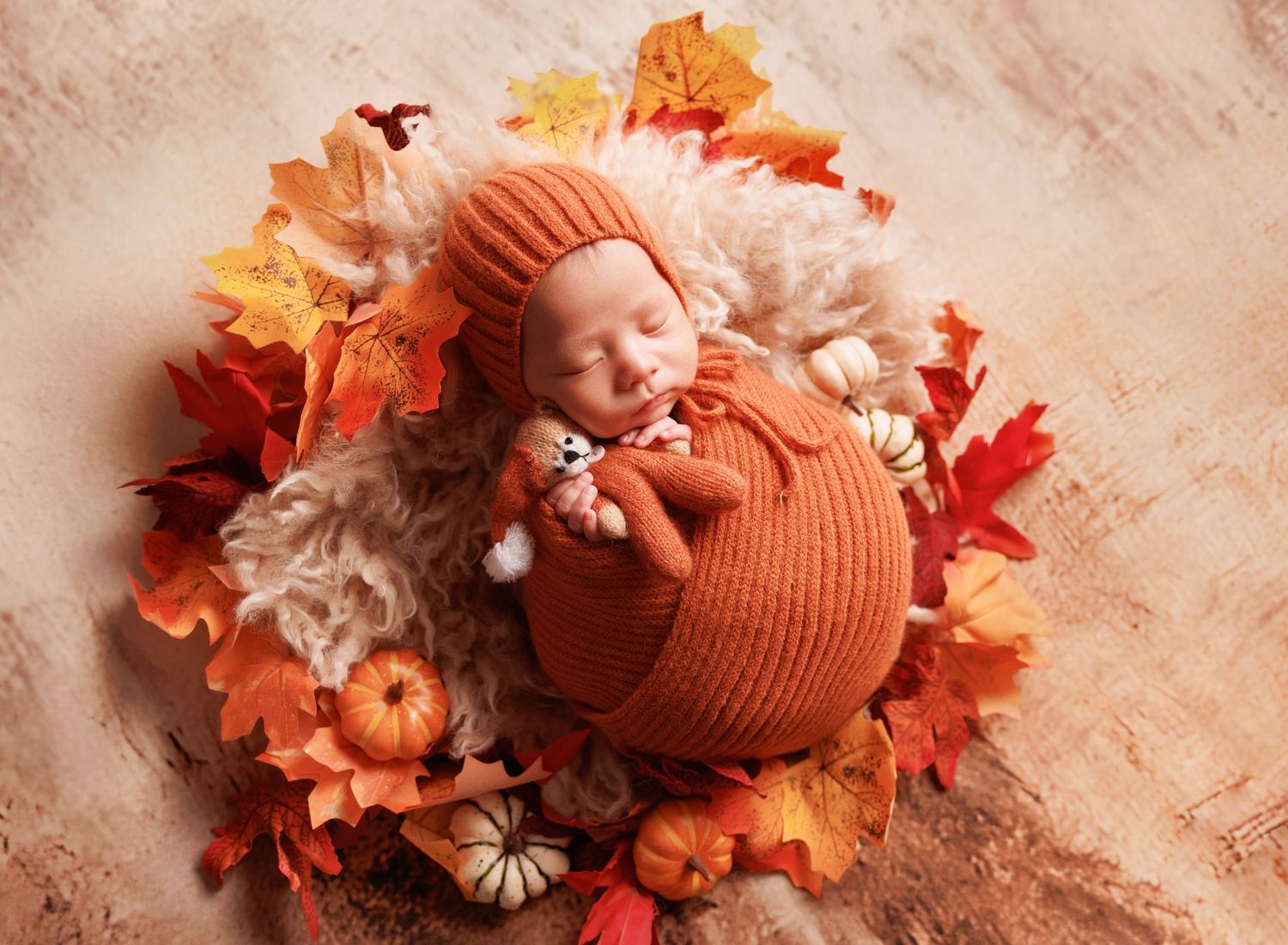 Newborn baby in orange knit romper and bonnet sleeping among colorful autumn leaves and small pumpkins.